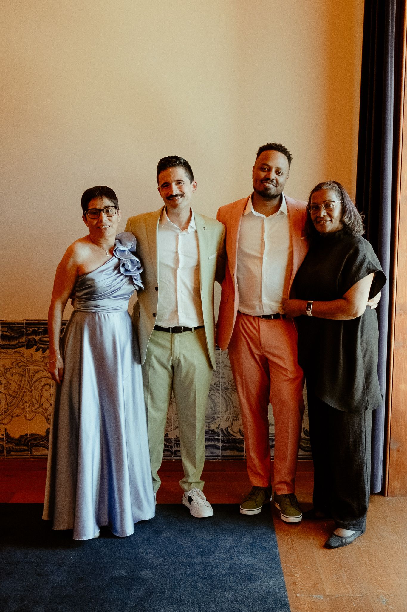 Four elegantly dressed people posing together at a formal event, wearing colorful outfits including blue gown and orange suit.