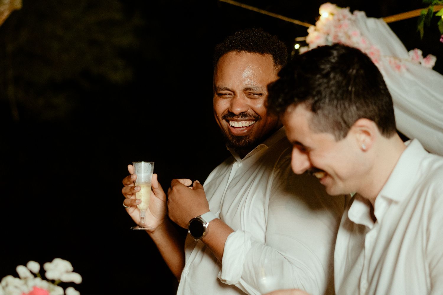 Two men laughing and celebrating at a wedding reception, one holding a drink under floral decorations at night.