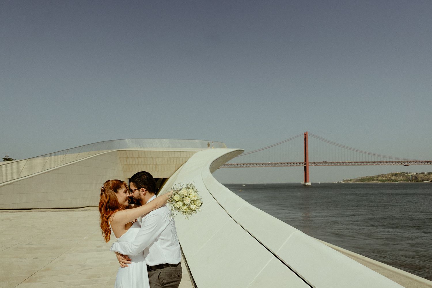Couple sharing a romantic kiss on a Lisbon waterfront with the 25 de Abril Bridge in the background.