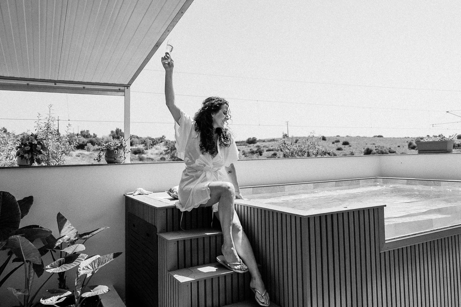 Woman in white dress sitting on rooftop terrace edge near hot tub, raising hand, black and white photo.