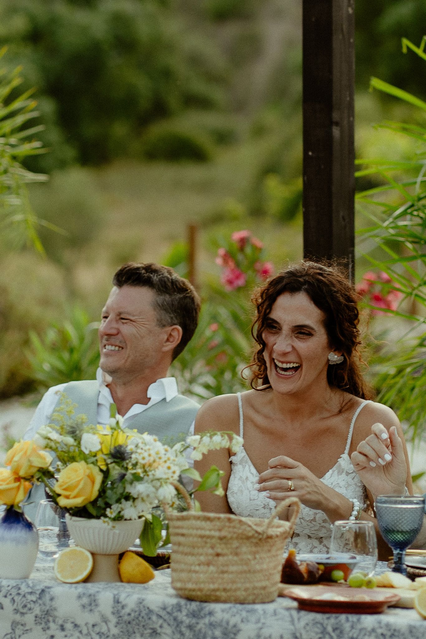 Laughing bride and groom seated at outdoor wedding reception table with yellow floral centerpiece and lush greenery.