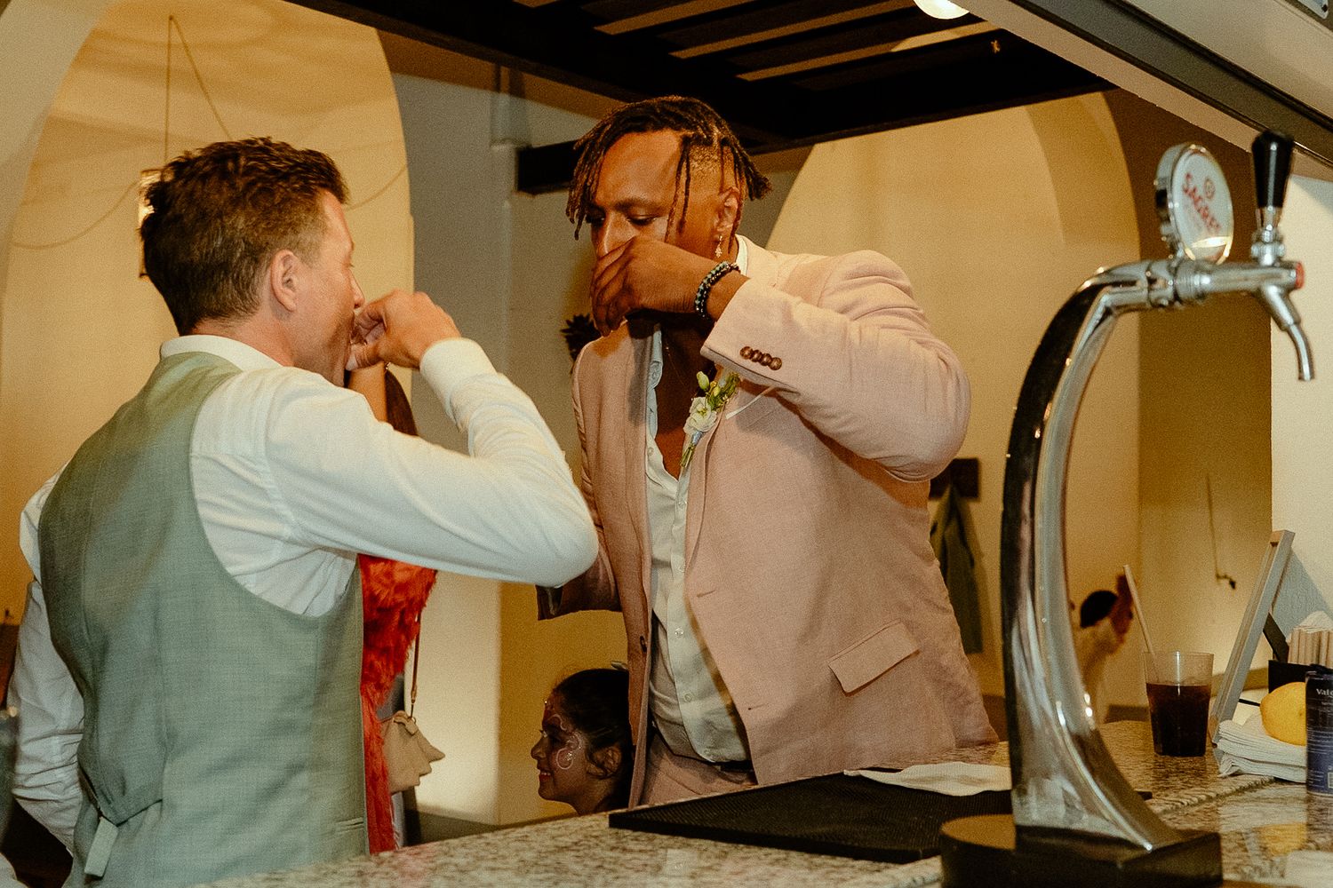 Two men in formal attire drinking shots at a bar counter with a beer tap visible nearby.