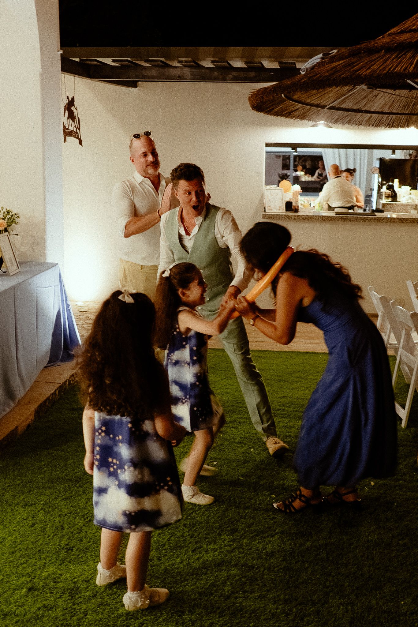 Adults and children playing together at an outdoor evening wedding reception on artificial grass.