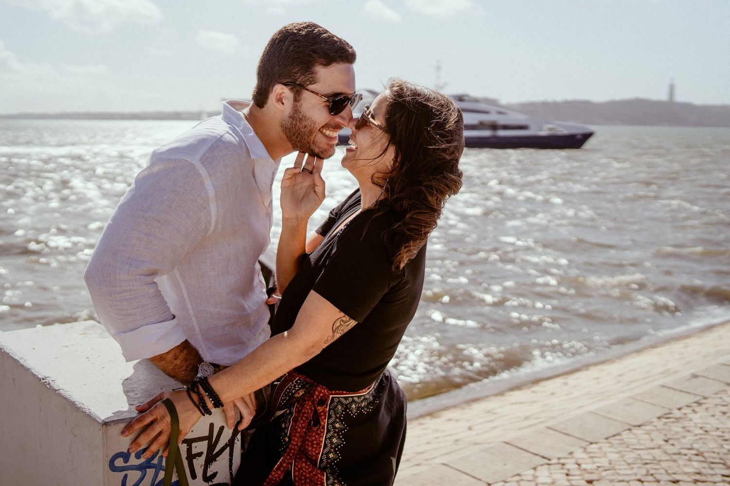 A romantic couple in casual beach attire embraces against a bright ocean backdrop during sunset.