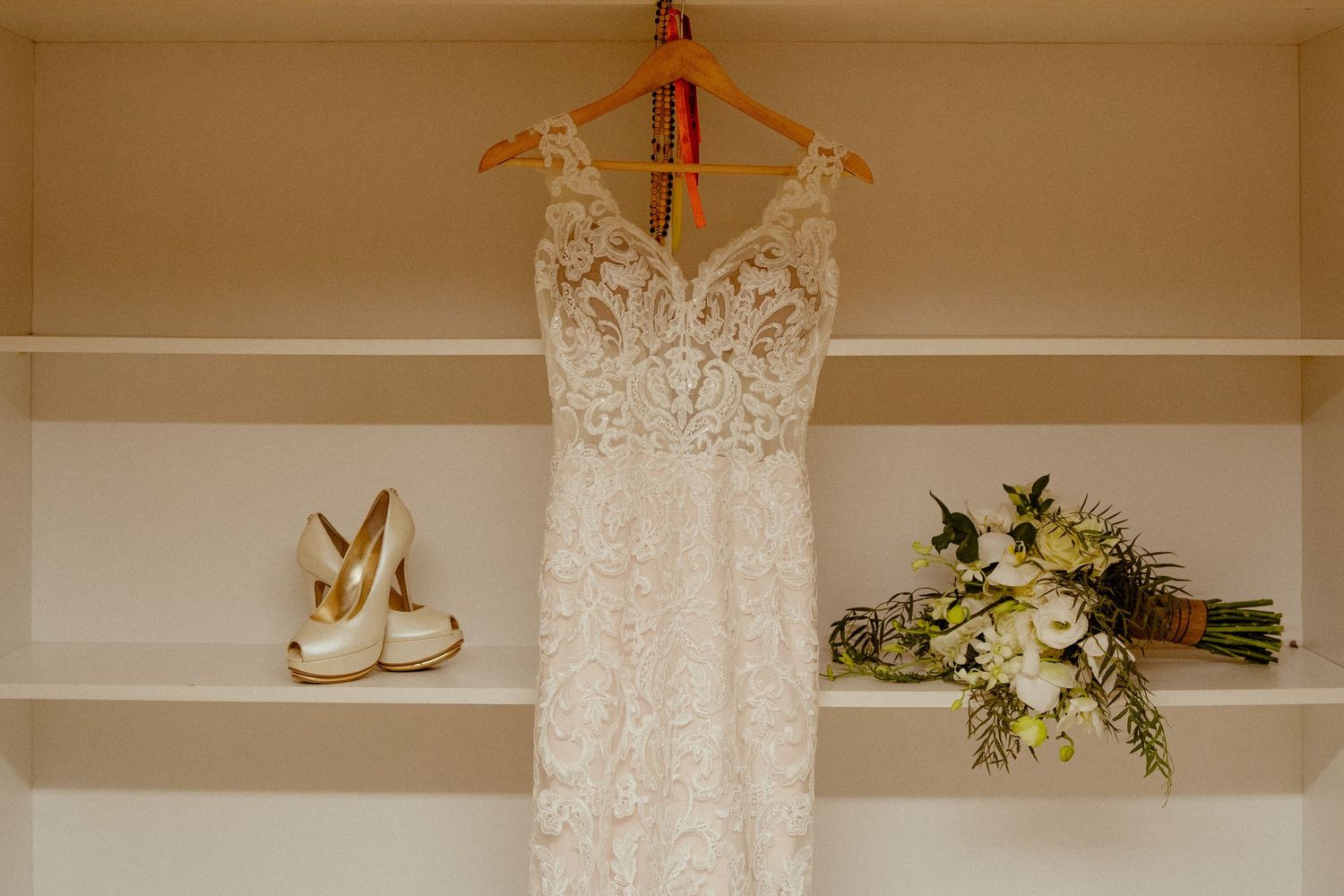 A delicate white lace wedding dress hangs beside ivory heels and a white floral bouquet on display shelves.