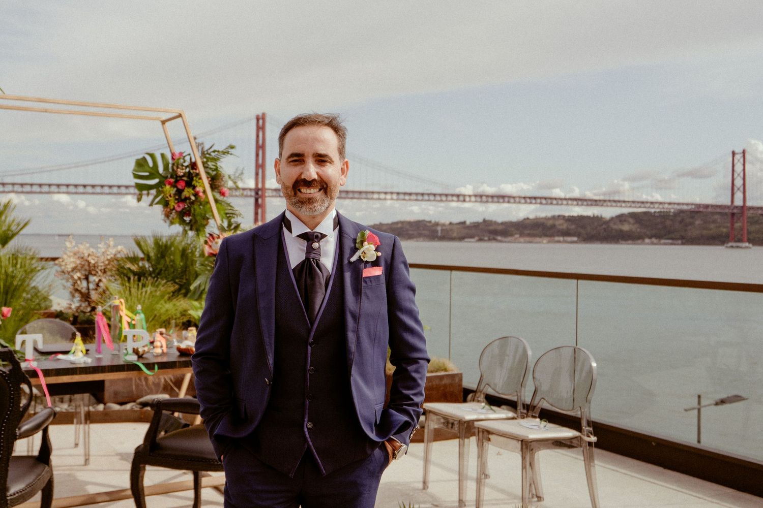 A person in a navy suit stands on a rooftop terrace overlooking the 25 de Abril Bridge in Lisbon, Portugal.