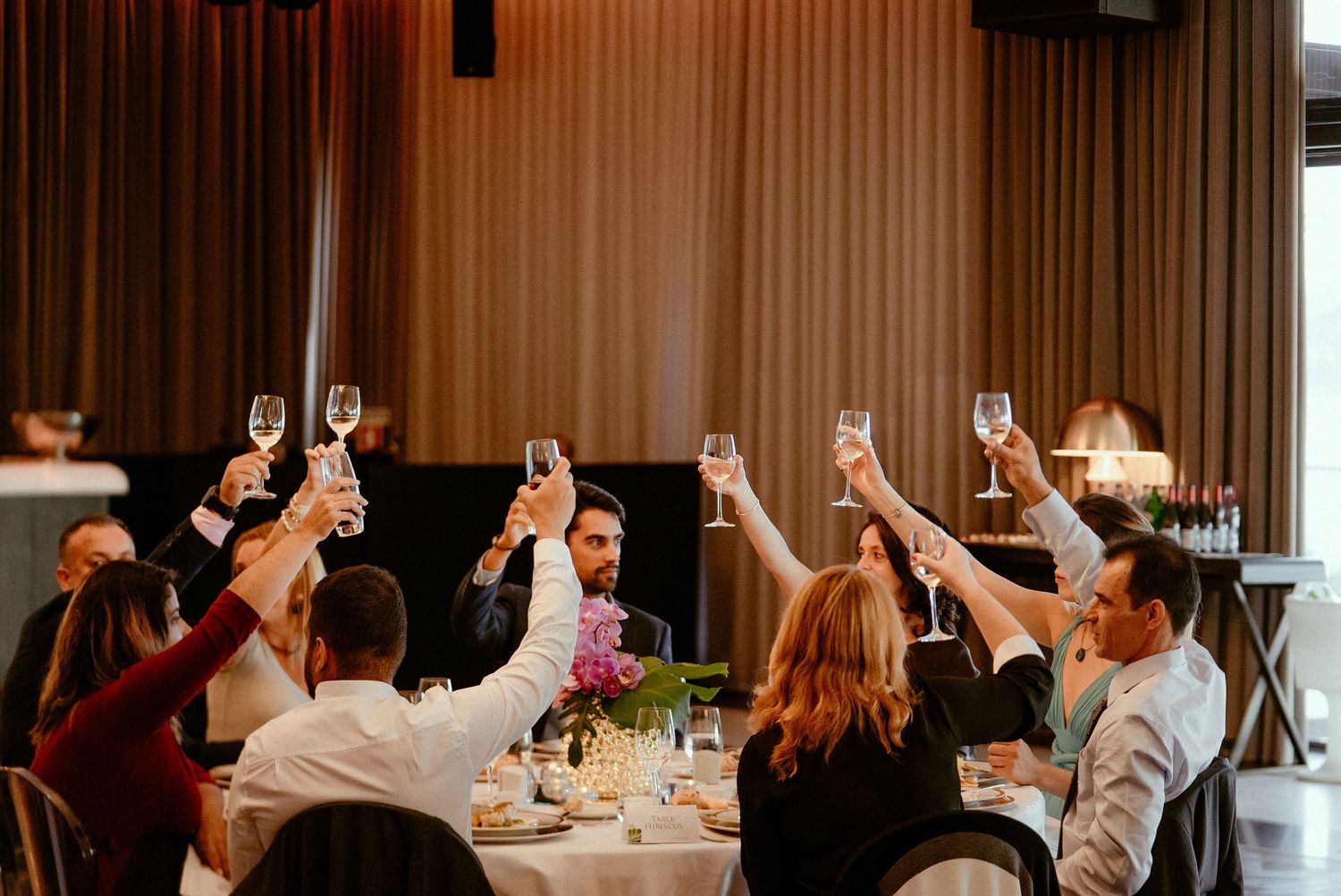 Wedding guests seated at tables raising their glasses for a celebratory toast at an indoor reception.