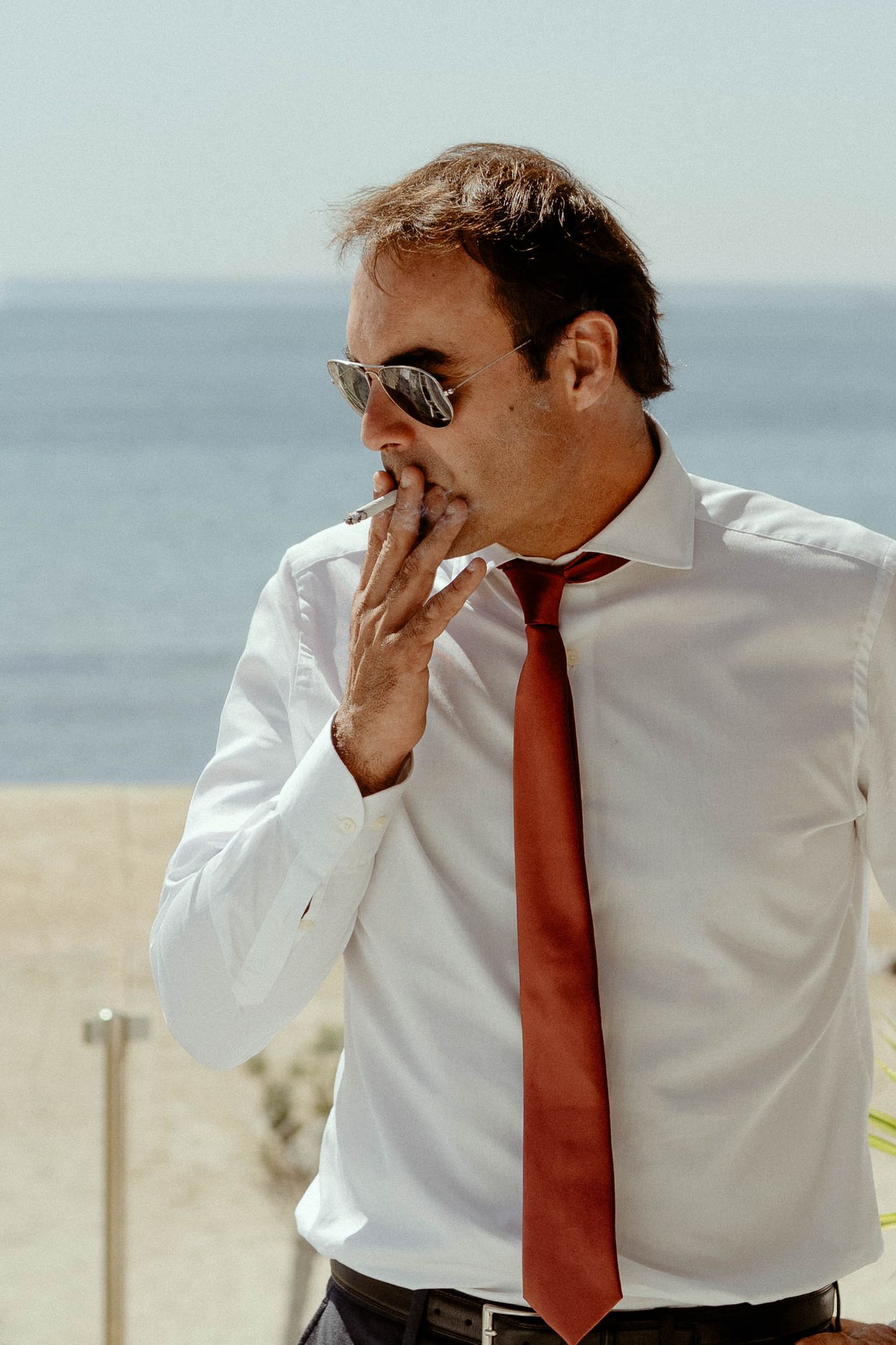Person in white dress shirt and red tie wearing sunglasses smoking while standing by the ocean on a sunny day.