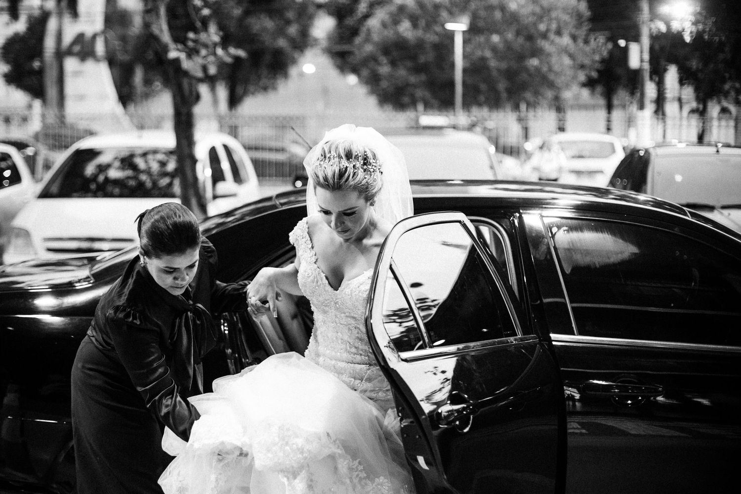 A wedding party assists a bride wearing a full white gown as she steps out of a black car.