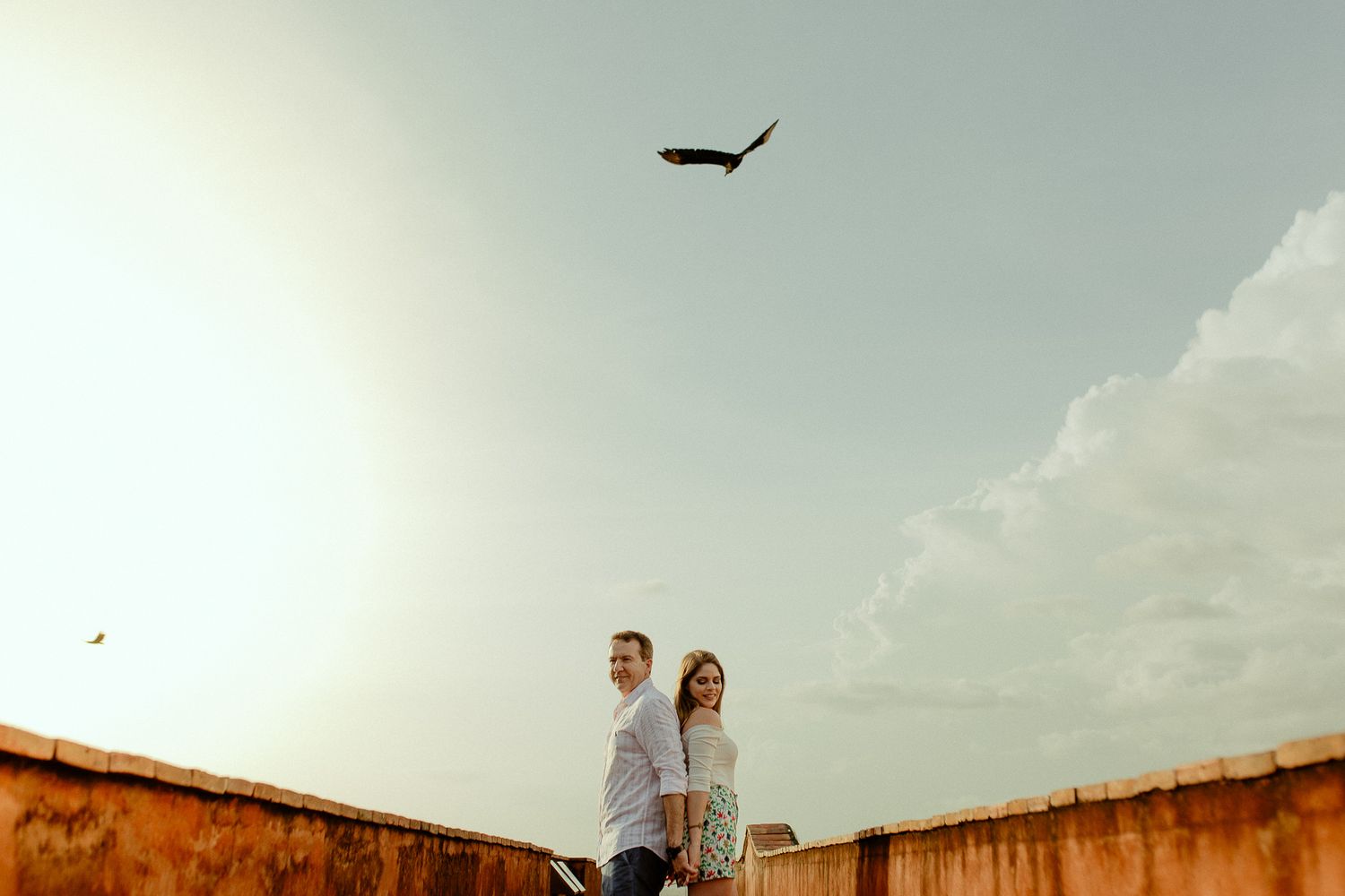 Two people stand on a rooftop against a cloudy sky with a bird soaring overhead.