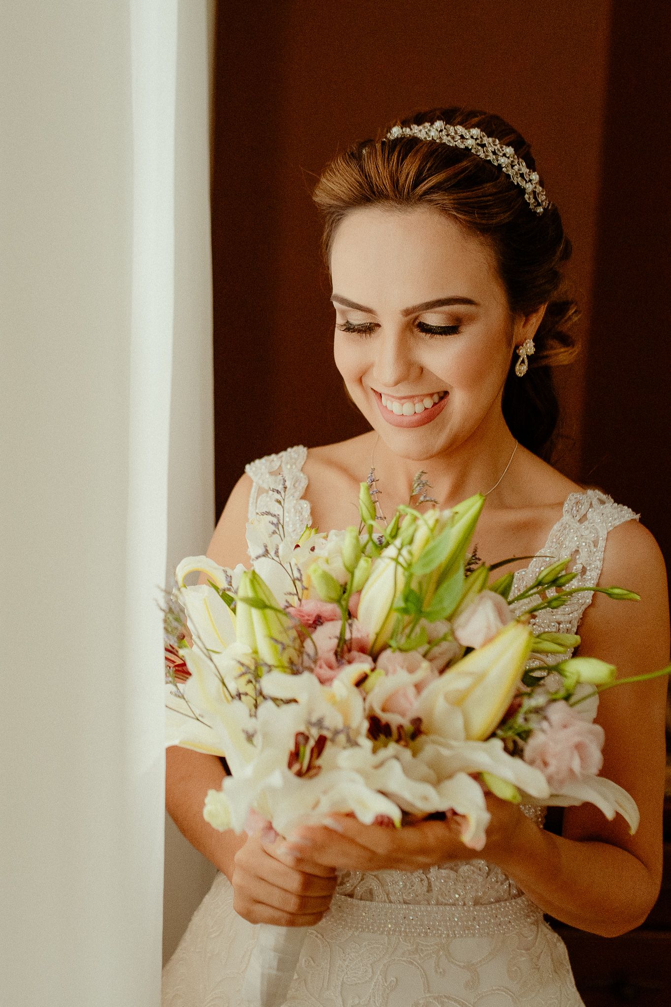 Bride in an elegant lace wedding dress holds a delicate bouquet of white lilies and pink flowers against a soft background.