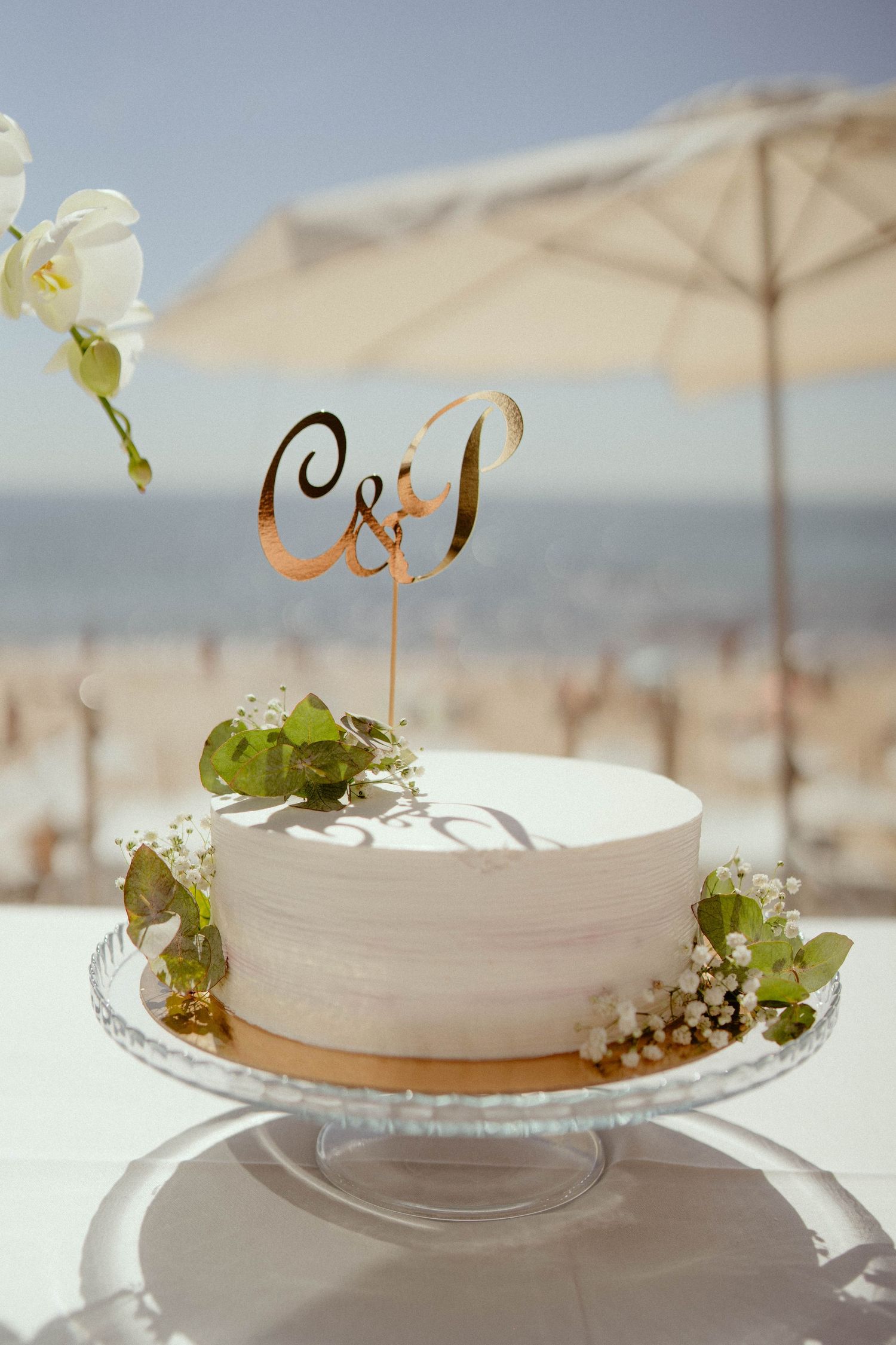 A white wedding cake decorated with delicate green foliage and a simple topper sits elegantly on a table under beach umbrellas.