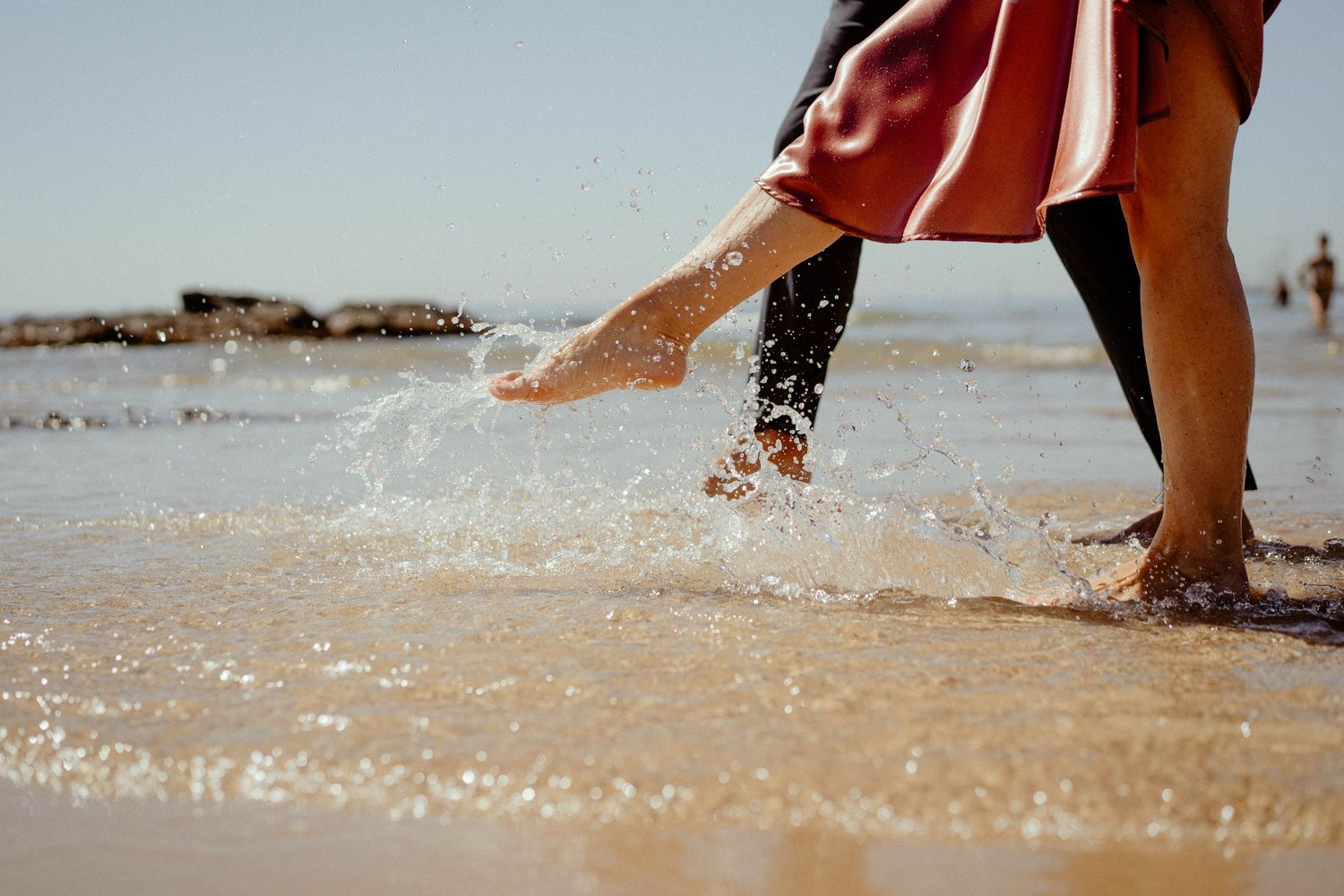 Bare feet splashing in shallow ocean waves on a sandy beach during a sunny summer day.