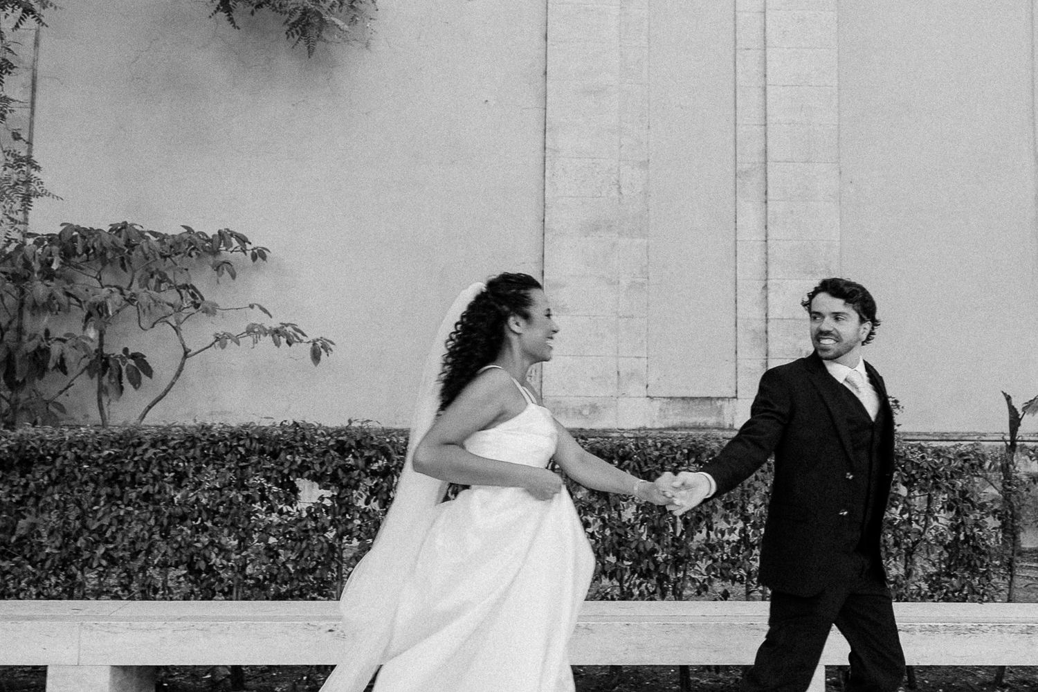 Black and white photo of a bride and groom holding hands, walking outdoors near a hedge and wall.