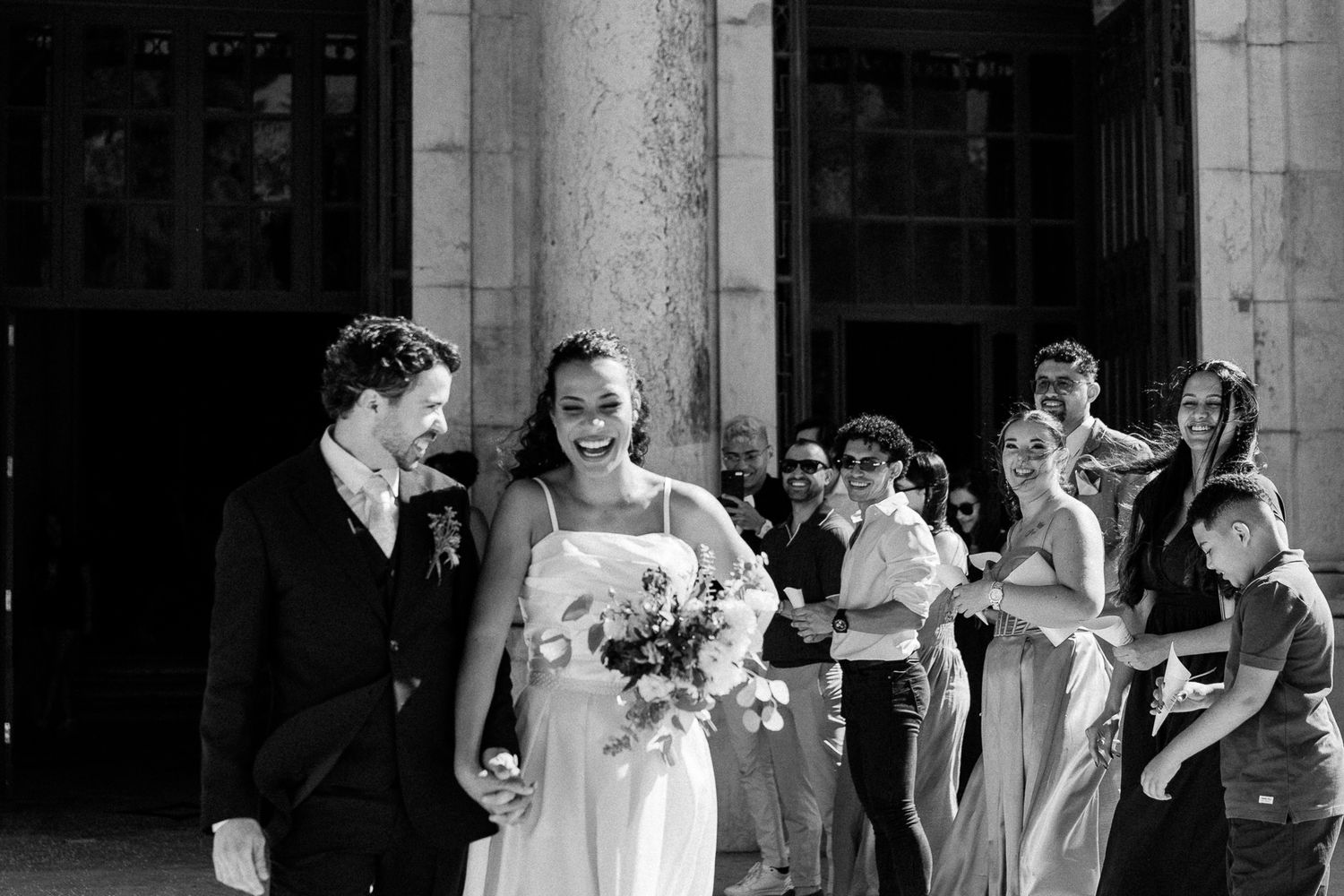 Black and white photo of a joyful bride holding bouquet walking with groom outside elegant building with guests.