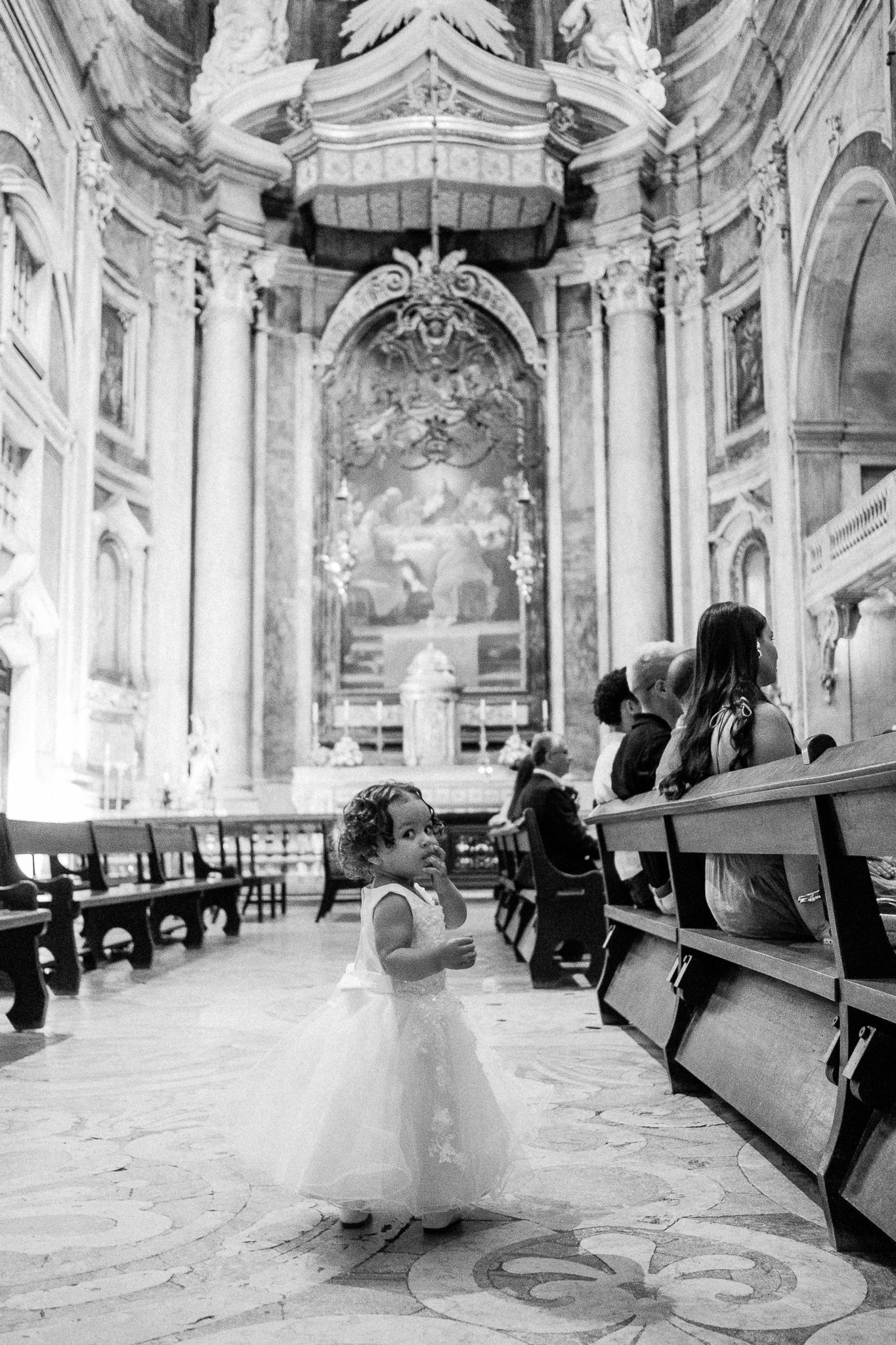 A little girl in a white dress stands in an ornate baroque church aisle during a wedding ceremony.