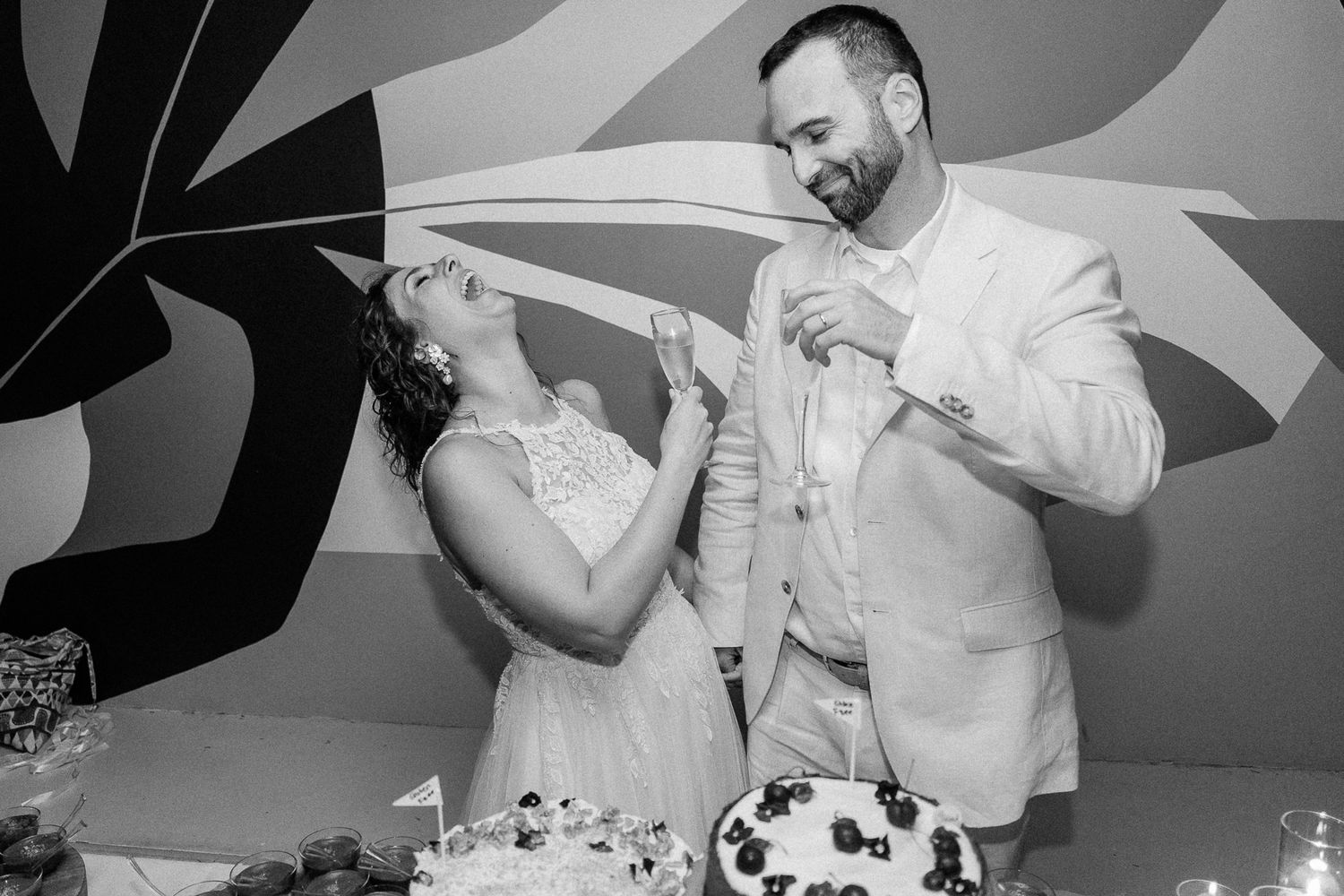 Black and white photo of a bride and groom laughing while cutting their wedding cake at their reception.
