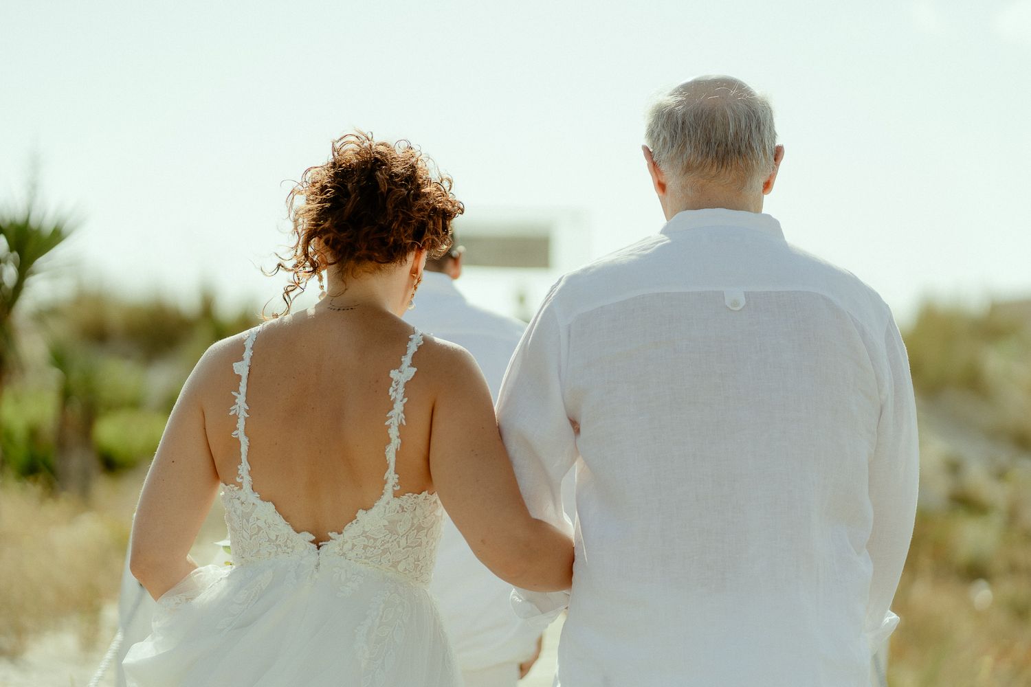 Bride and groom walking together outdoors in white wedding attire on a sunny day.