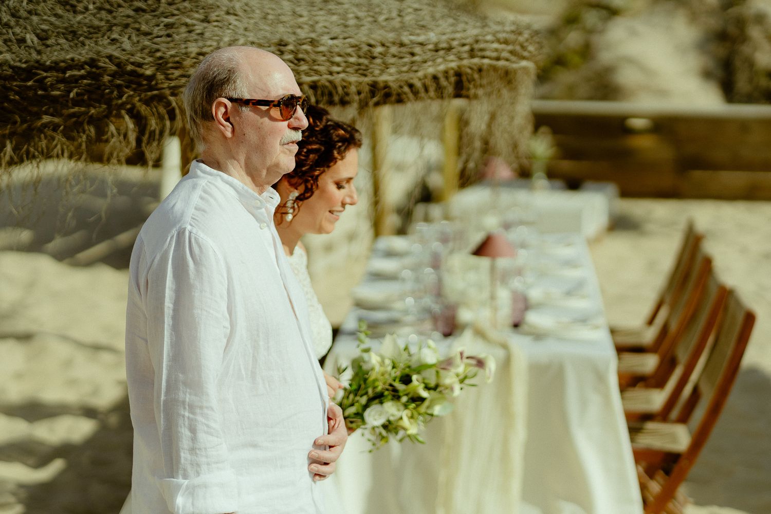 Couple in white attire stands together on a beach near an elegant outdoor dining table setup.