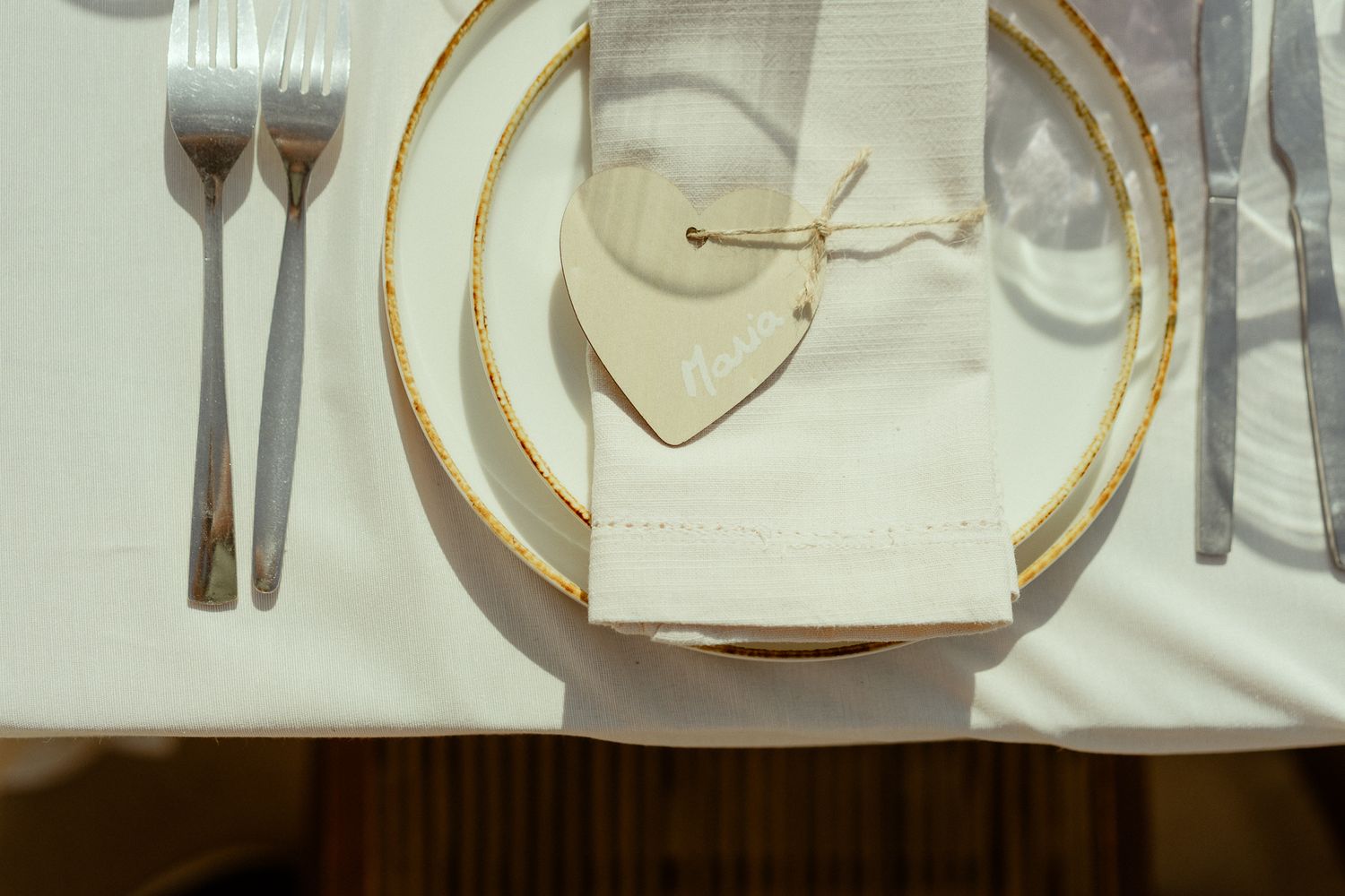 Elegant gold-rimmed place setting with white napkin, heart-shaped tag, and silverware on white tablecloth.