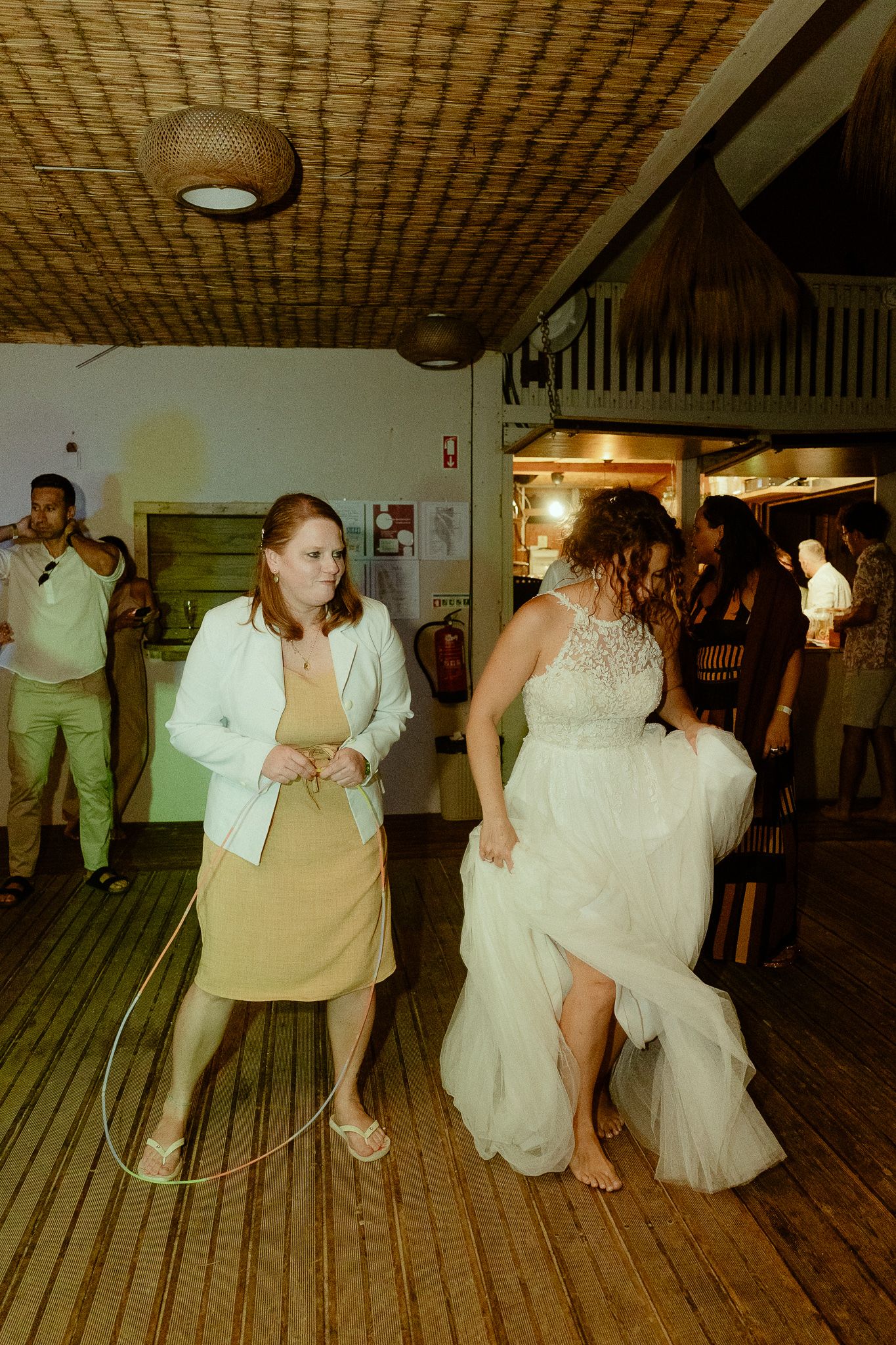 Bride in white gown and guest in yellow dress hula hooping at a rustic wedding reception venue.