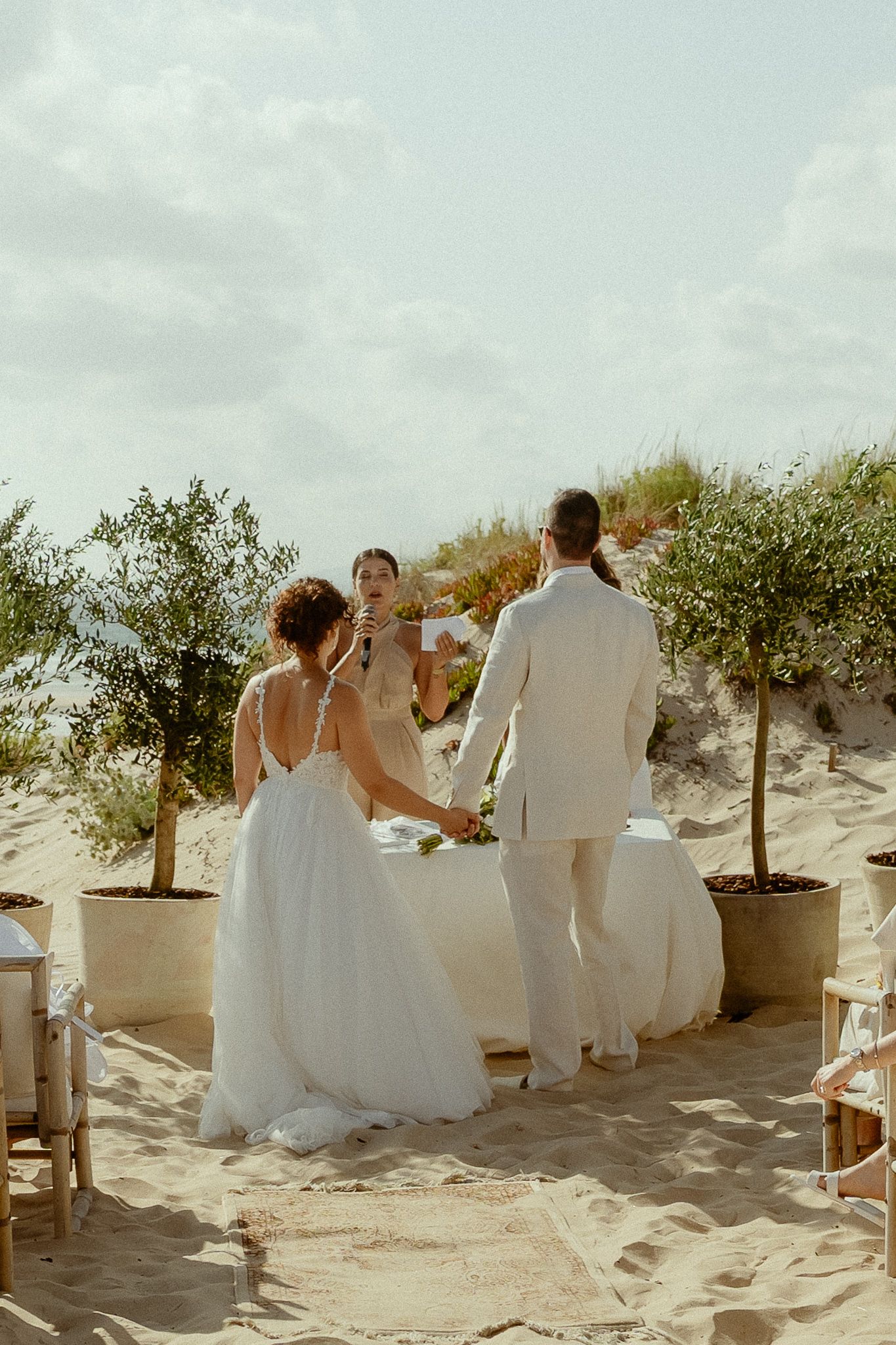 Bride and groom exchange vows at a romantic beach wedding ceremony with potted olive trees on sandy dunes.
