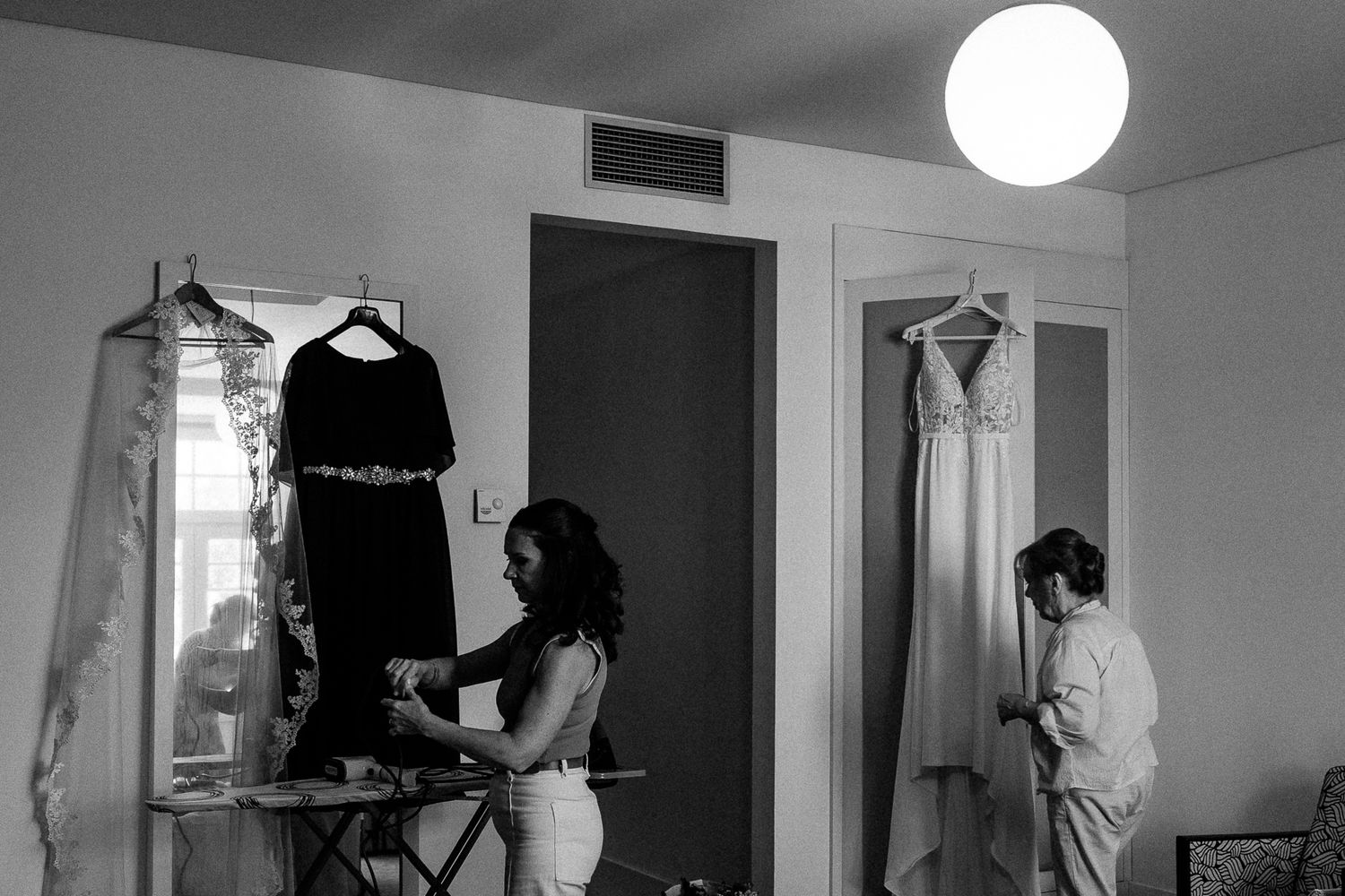 Black and white photo of bride ironing dress while bridesmaid adjusts gown on hanger in elegant wedding preparation room.