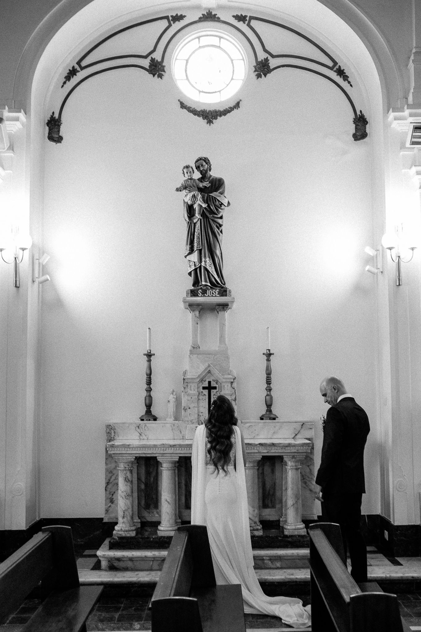 Black and white photo of a bride and groom standing before a church altar with a saint statue during their wedding ceremony.