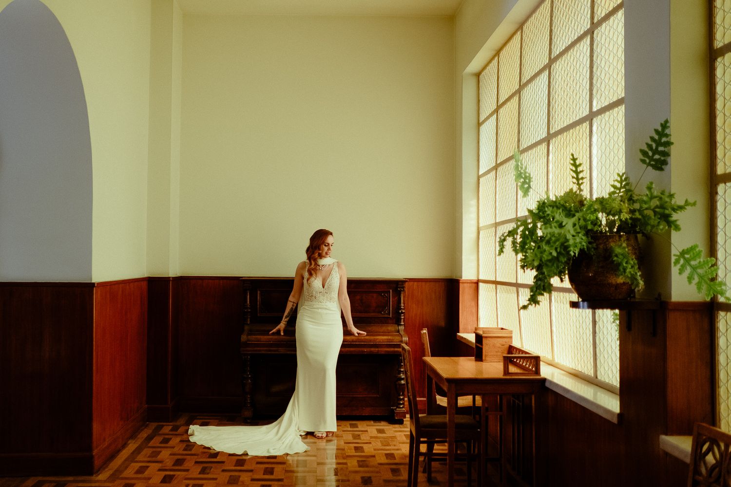 Bride in fitted white gown stands by sunlit windows near vintage piano in warm interior.