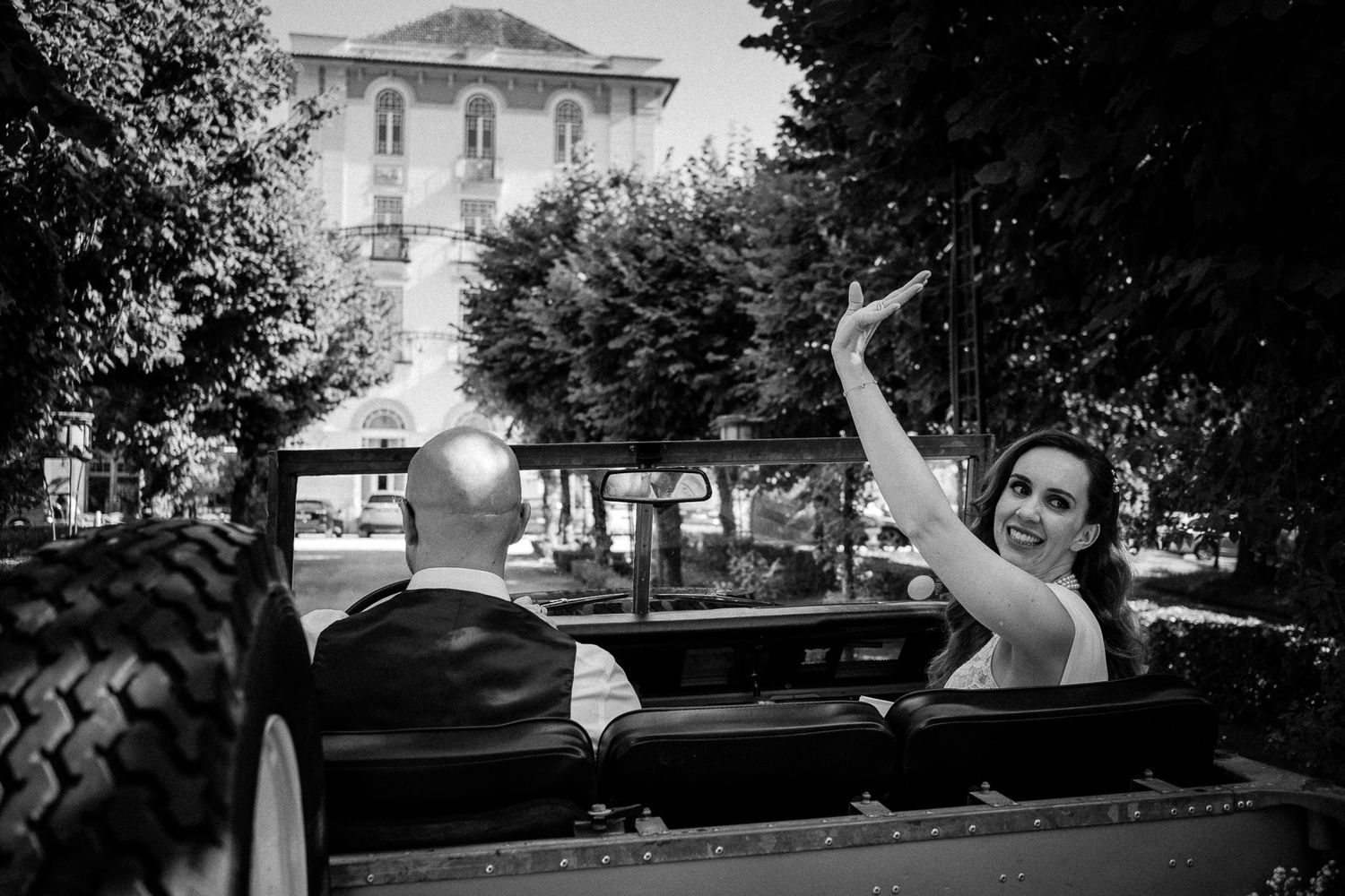 Bride and groom ride in vintage convertible, bride waving, elegant black and white photo.