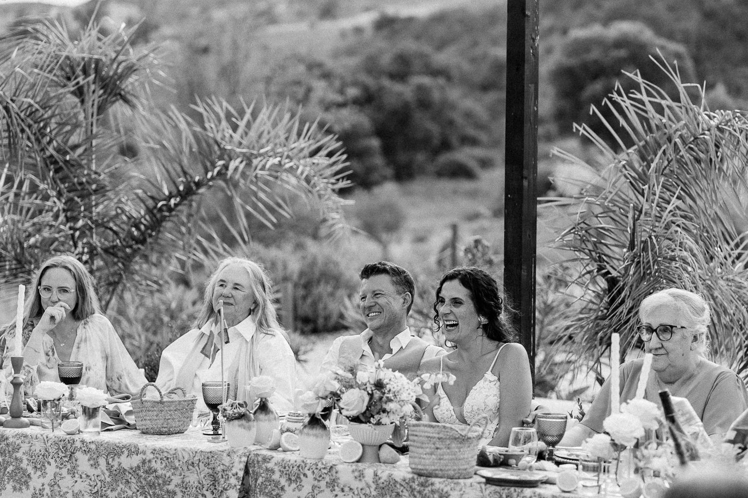 Black and white wedding reception photo of bride, groom, and guests laughing at outdoor table with tropical palms.