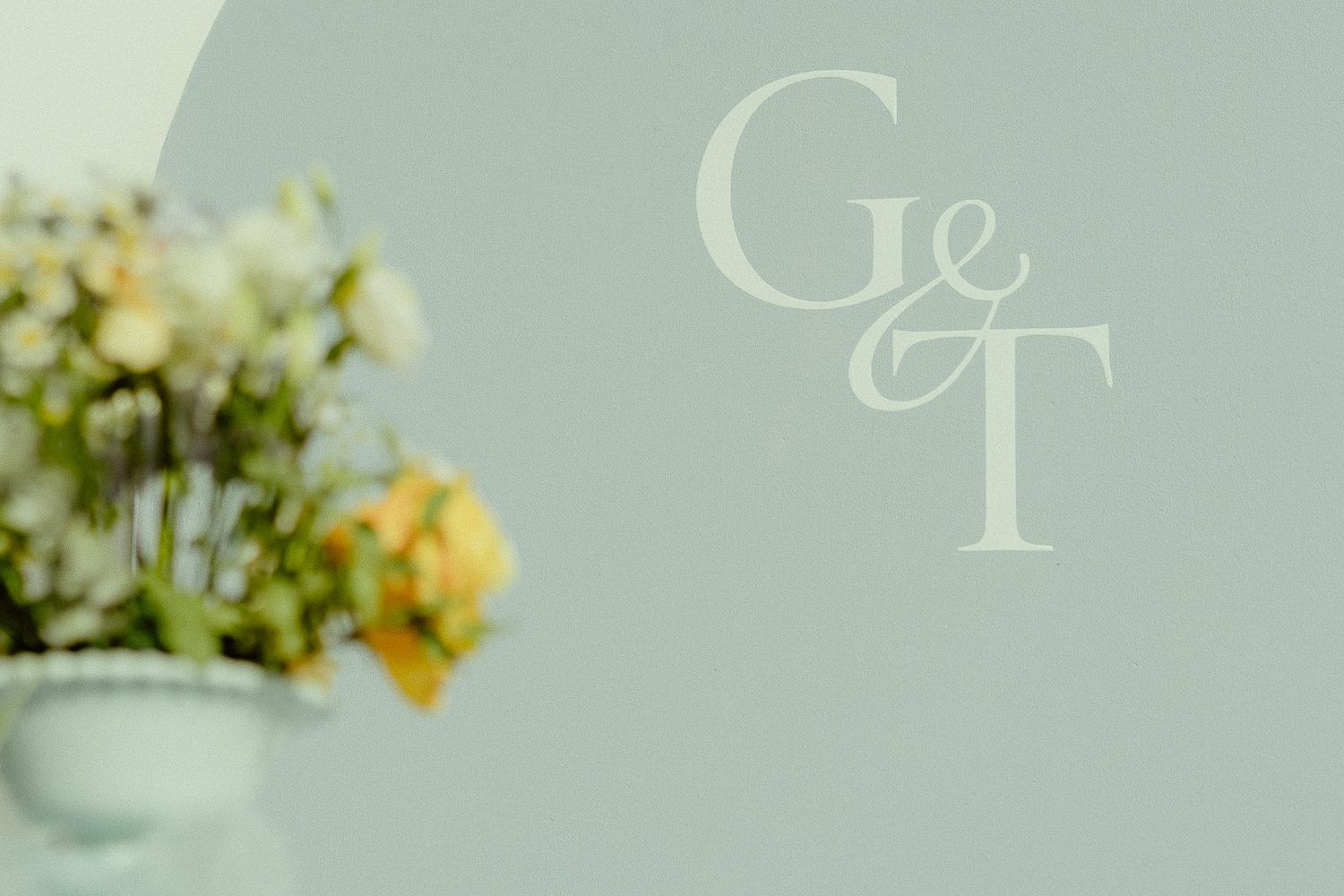 Elegant G&T monogram logo on a soft sage green wall with blurred floral arrangement in foreground.