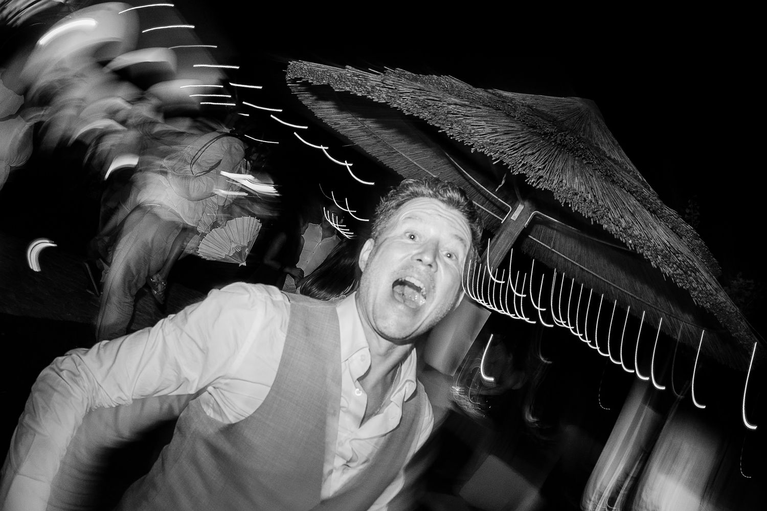 Black and white photo of a man dancing energetically at an outdoor party with motion blur effect.