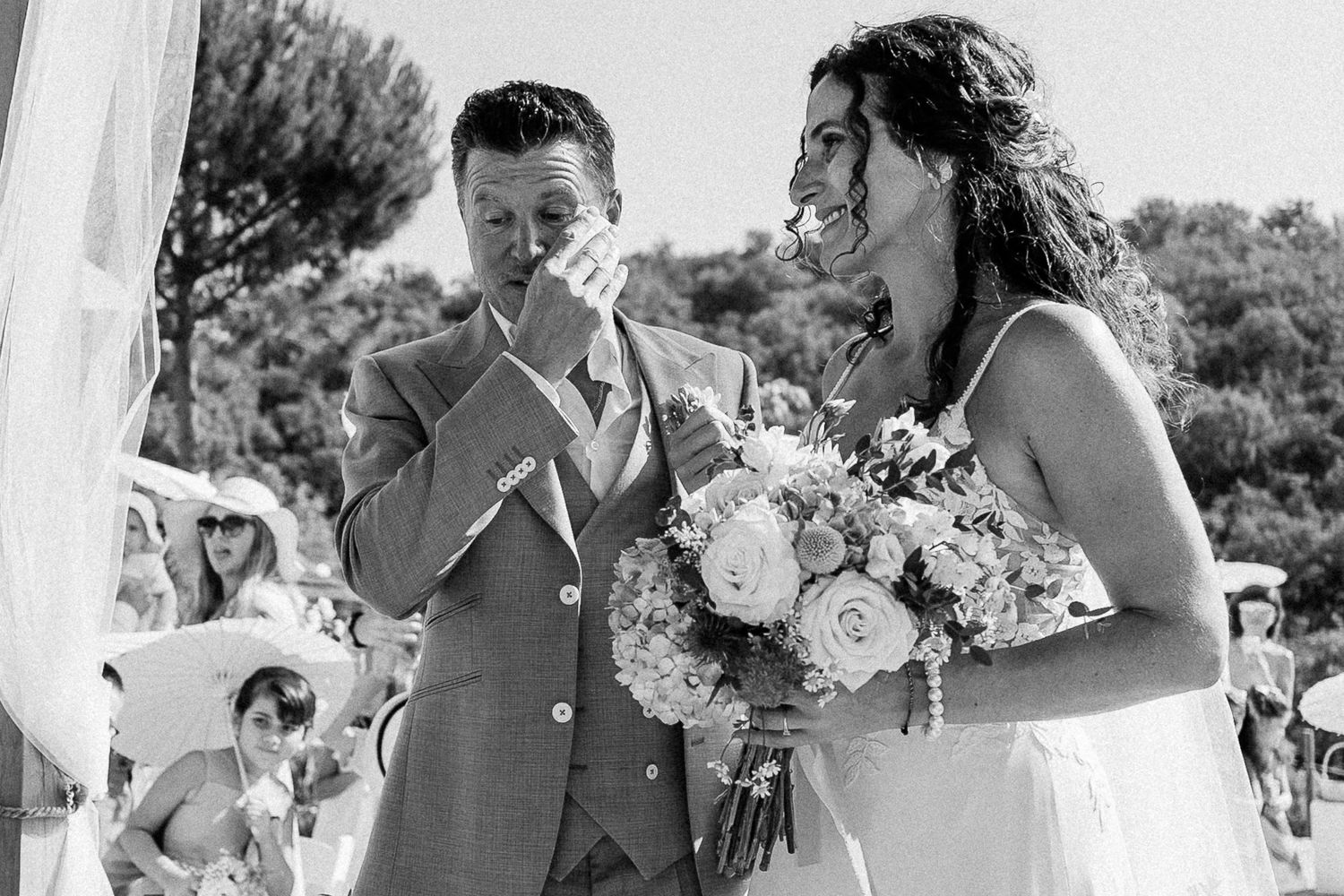 Emotional groom wiping tears beside smiling bride holding bouquet at outdoor wedding ceremony in black and white photo.