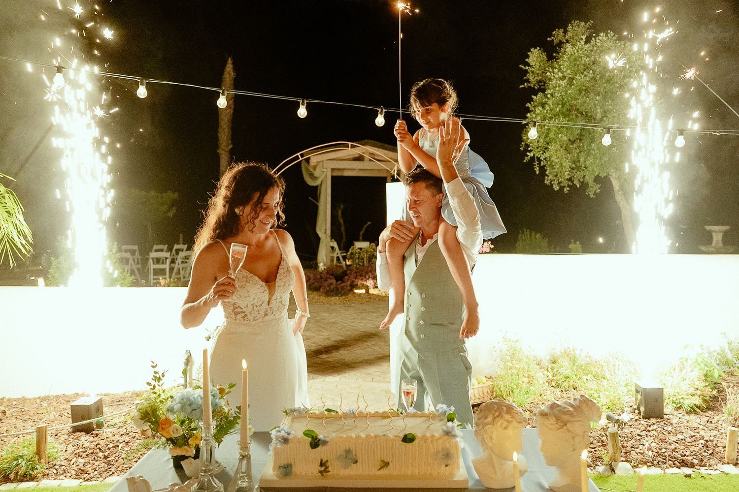 Couple celebrating wedding with child and sparklers beside tiered cake at outdoor night reception.