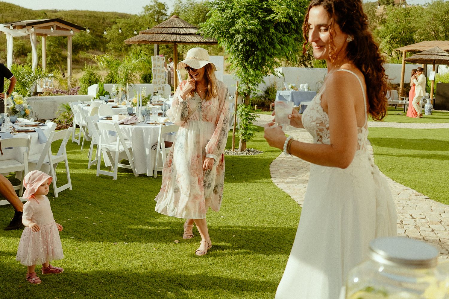 Elegant outdoor wedding reception with guests in white and pink dresses on green lawn with decorated tables.