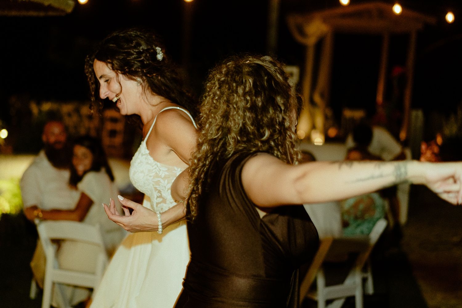 Two women dancing joyfully at an outdoor wedding reception at night, one in a white bridal gown, guests seated in background.