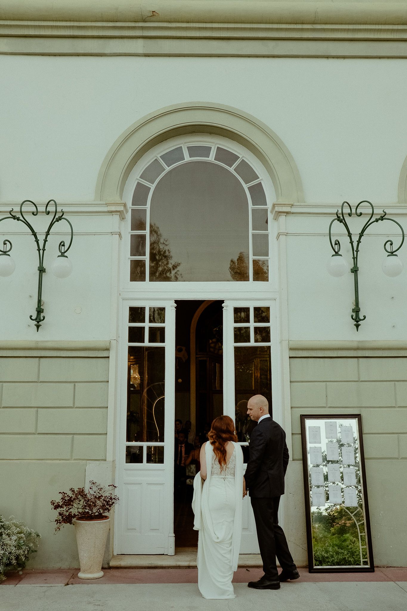 Bride and groom stand before elegant white arched doorway of wedding venue, bride in flowing white gown.