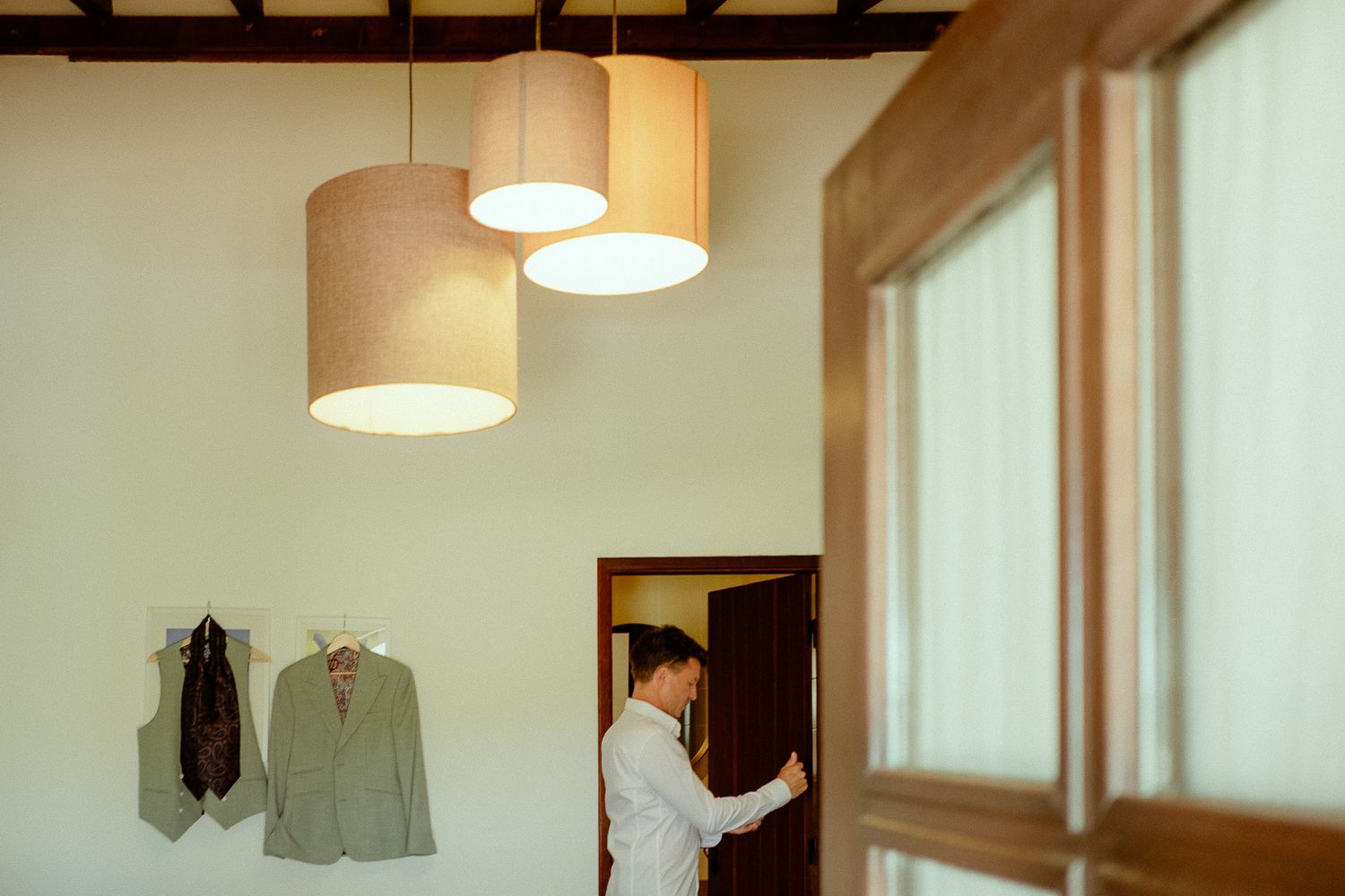 Groom preparing in elegant room with hanging pendant lights and suit displayed on wall hooks.