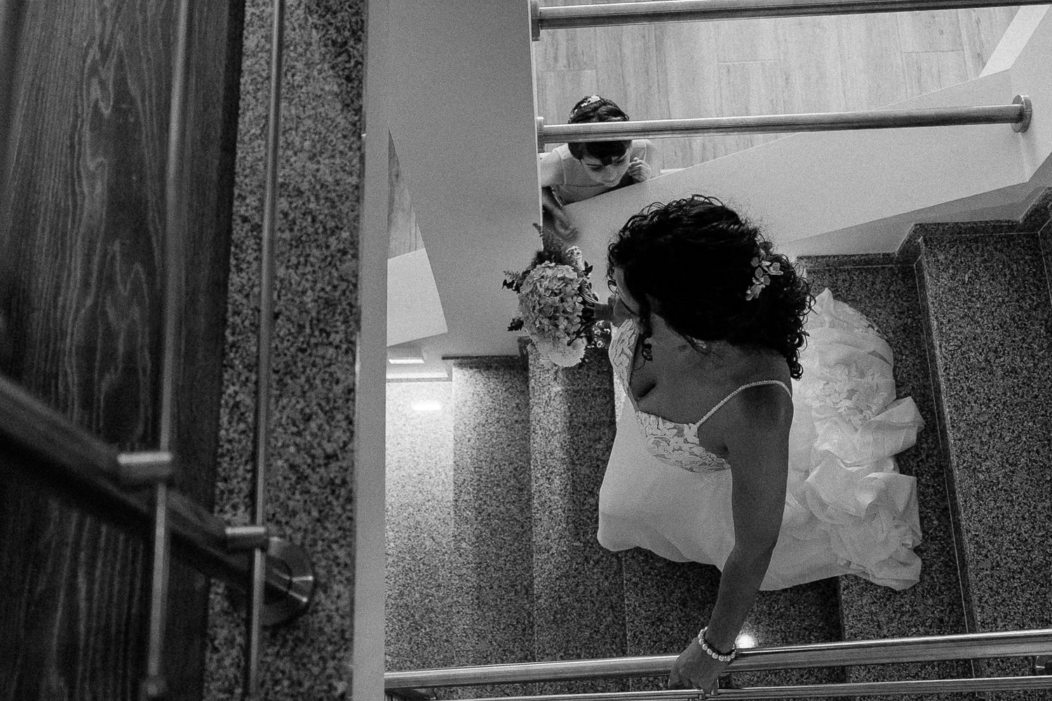 Black and white photo of a bride in a white gown holding a bouquet, descending elegant stairs on her wedding day.