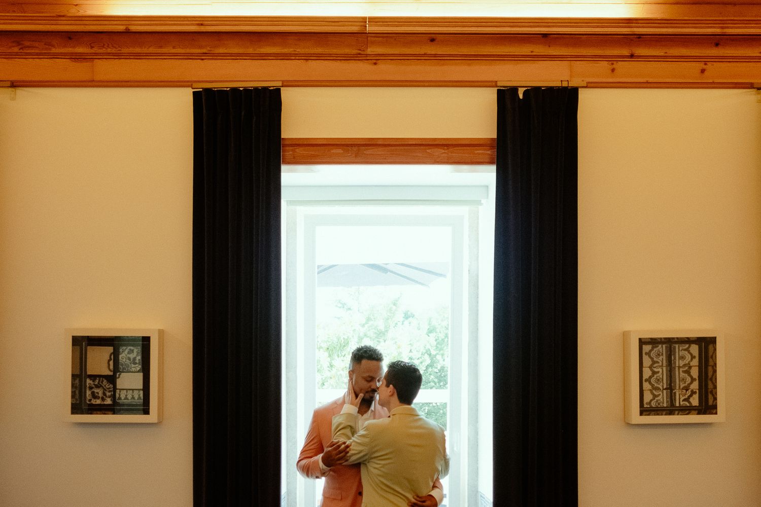Two men share a romantic dance by a sunlit window, framed by dark curtains and artwork on white walls.