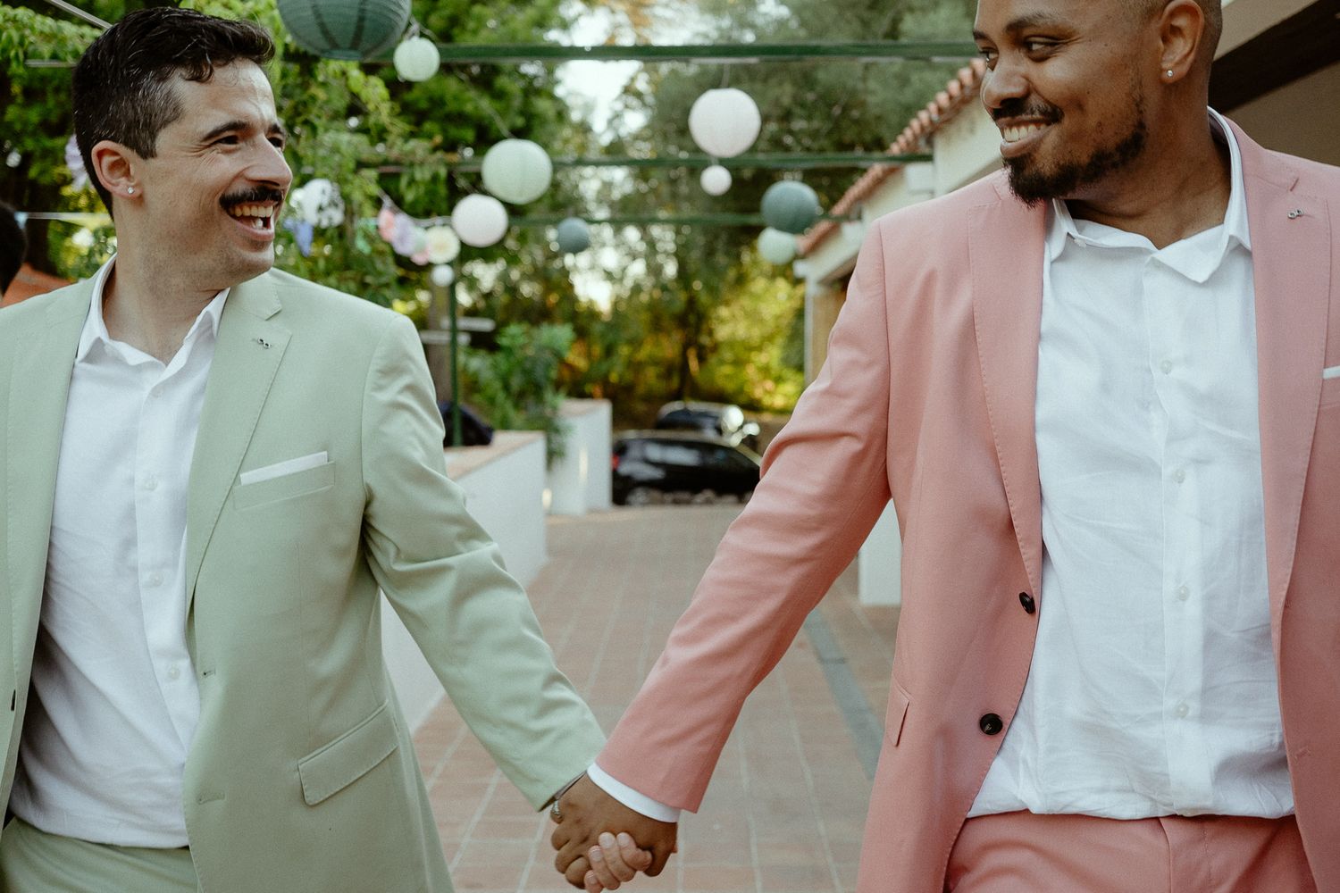 Two grooms in pastel suits holding hands and smiling joyfully at their outdoor wedding celebration.