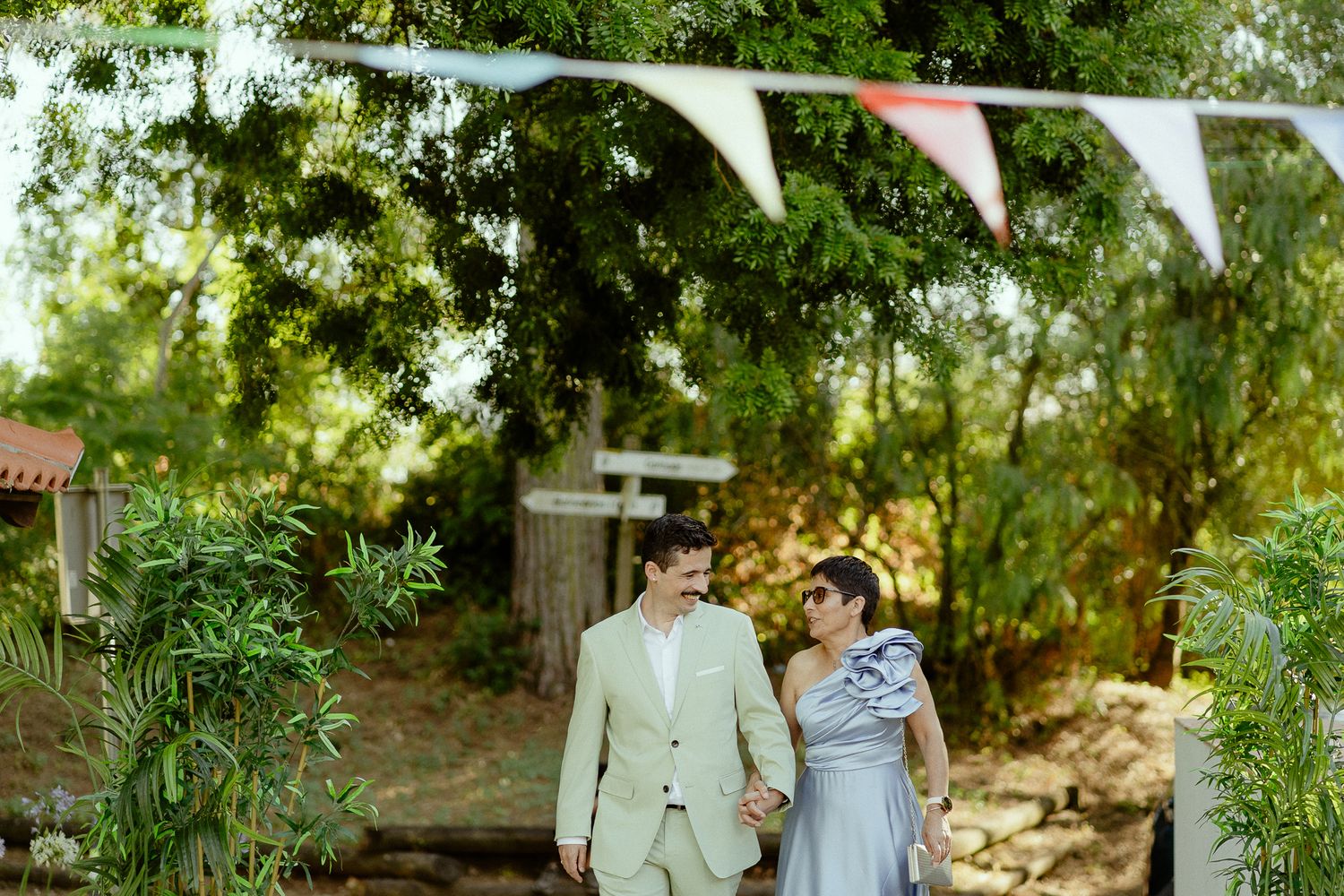 Couple holding hands in lush garden setting with colorful bunting flags, man in beige suit, woman in blue dress.