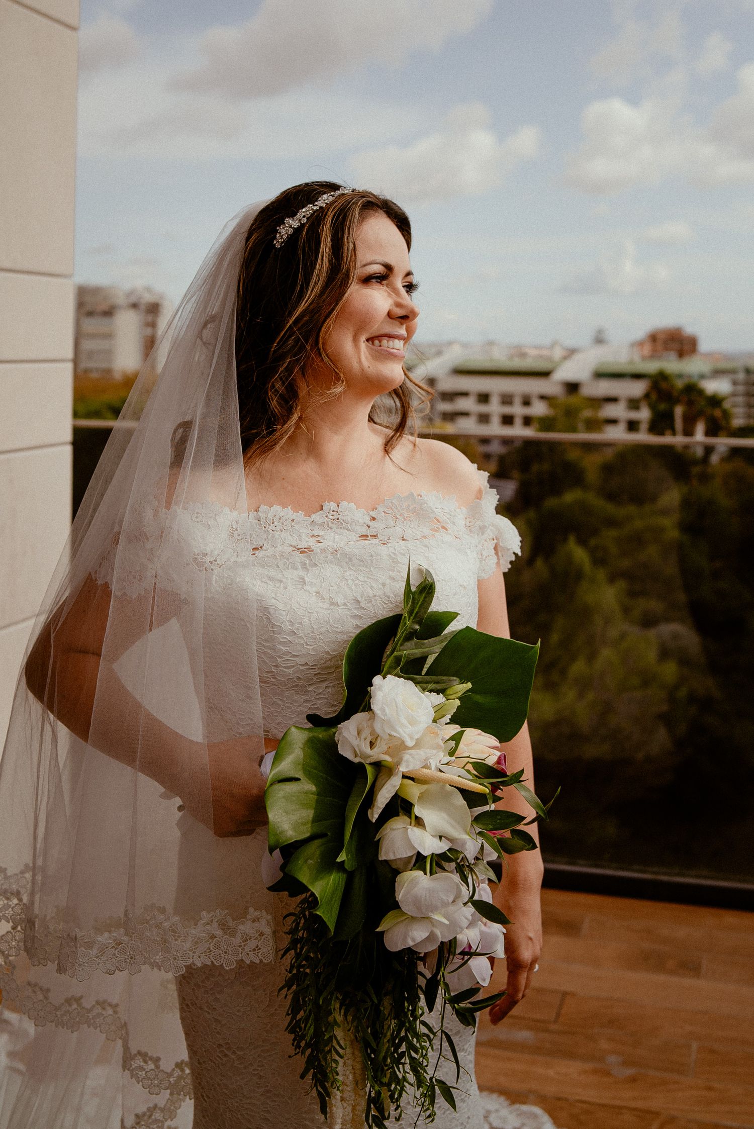 Bride in lace wedding dress holds white floral bouquet and looks into distance from outdoor balcony at sunset.