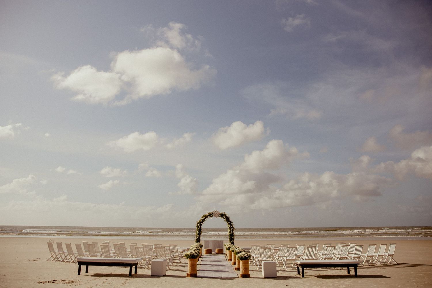 Wedding ceremony setup on a beach with white chairs and wooden arch under cloudy skies.