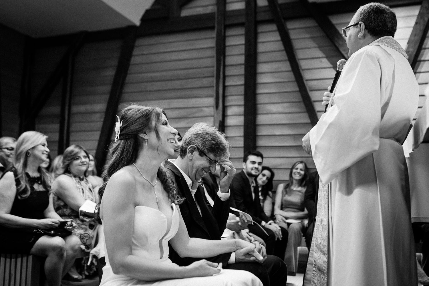 Black and white photo of a wedding ceremony in progress with people seated in chairs and a priest in white robes.