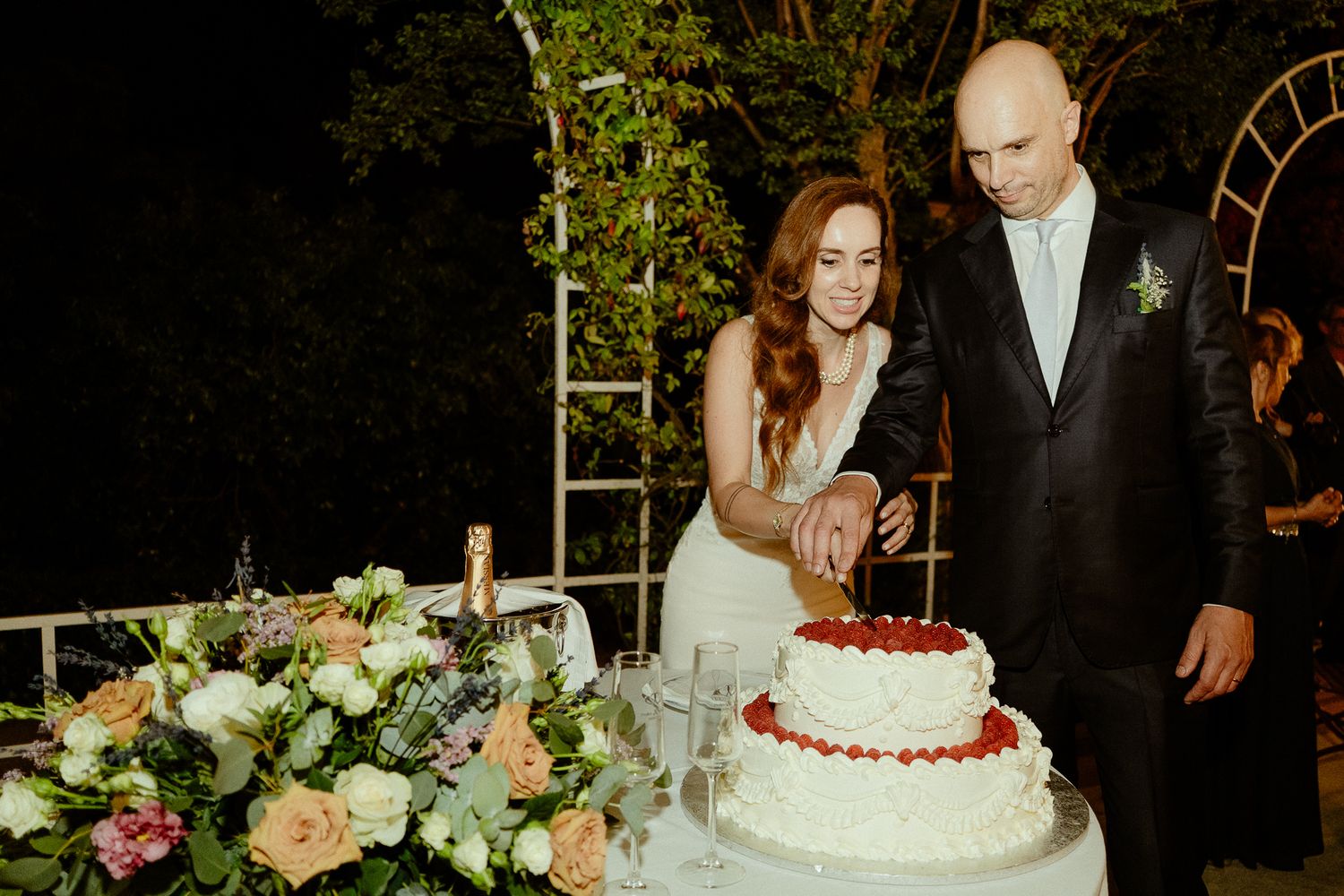 Bride and groom cutting a two-tier white wedding cake decorated with strawberries at an outdoor evening reception.