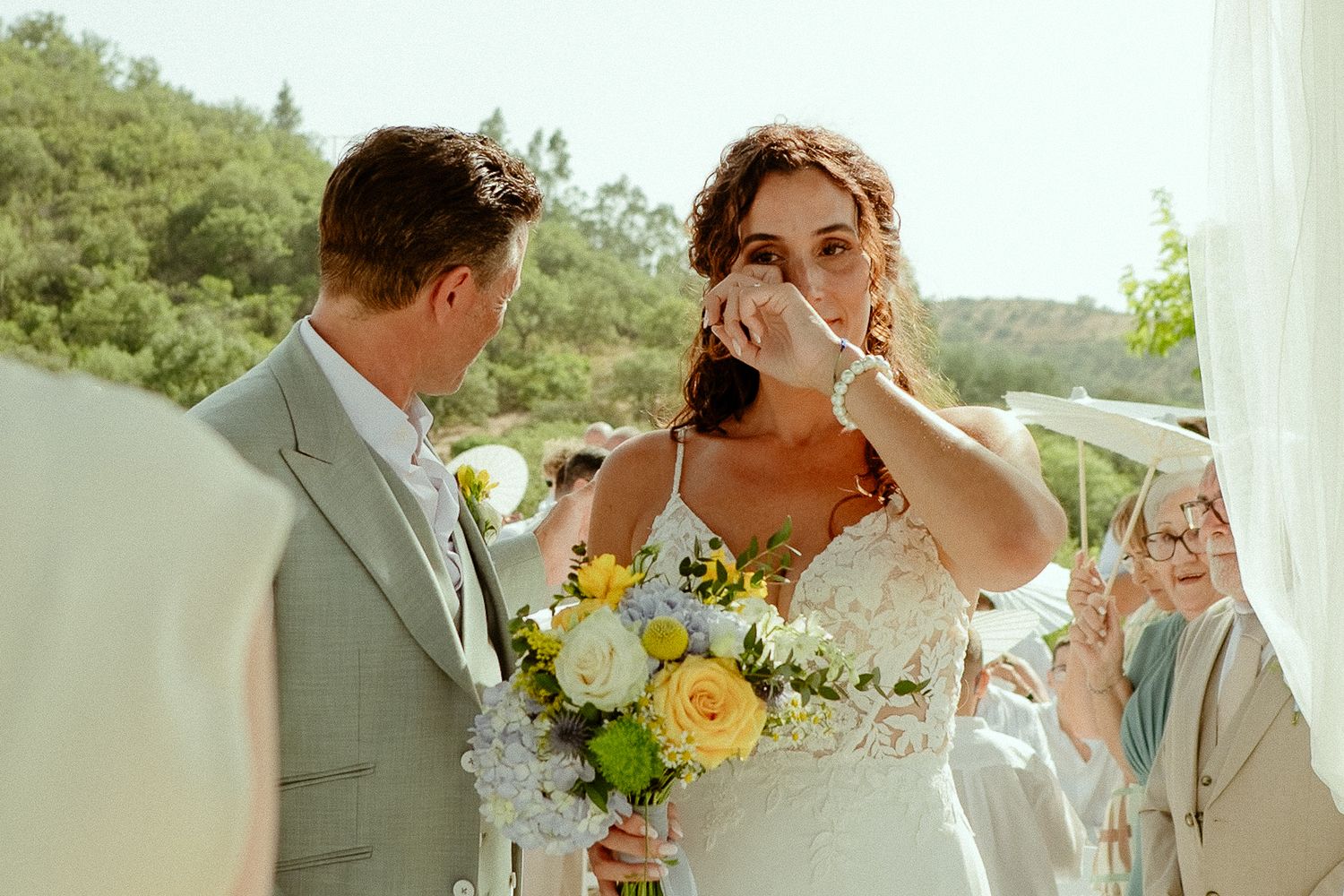 Emotional bride wiping tears holding yellow floral bouquet beside groom in grey suit at outdoor wedding ceremony.