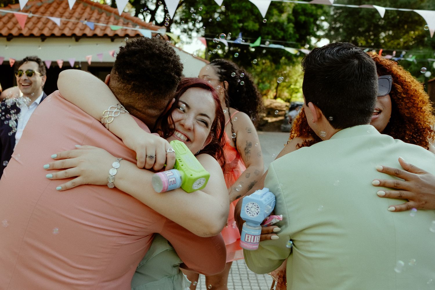 Joyful group hug at an outdoor celebration, with a smiling woman embracing friends amid bubbles and festive decor.