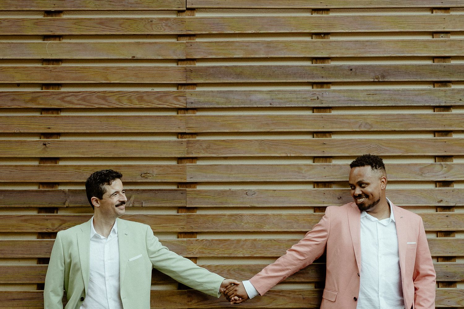 Two men in pastel suits hold hands and smile at each other against a wooden slat wall backdrop.